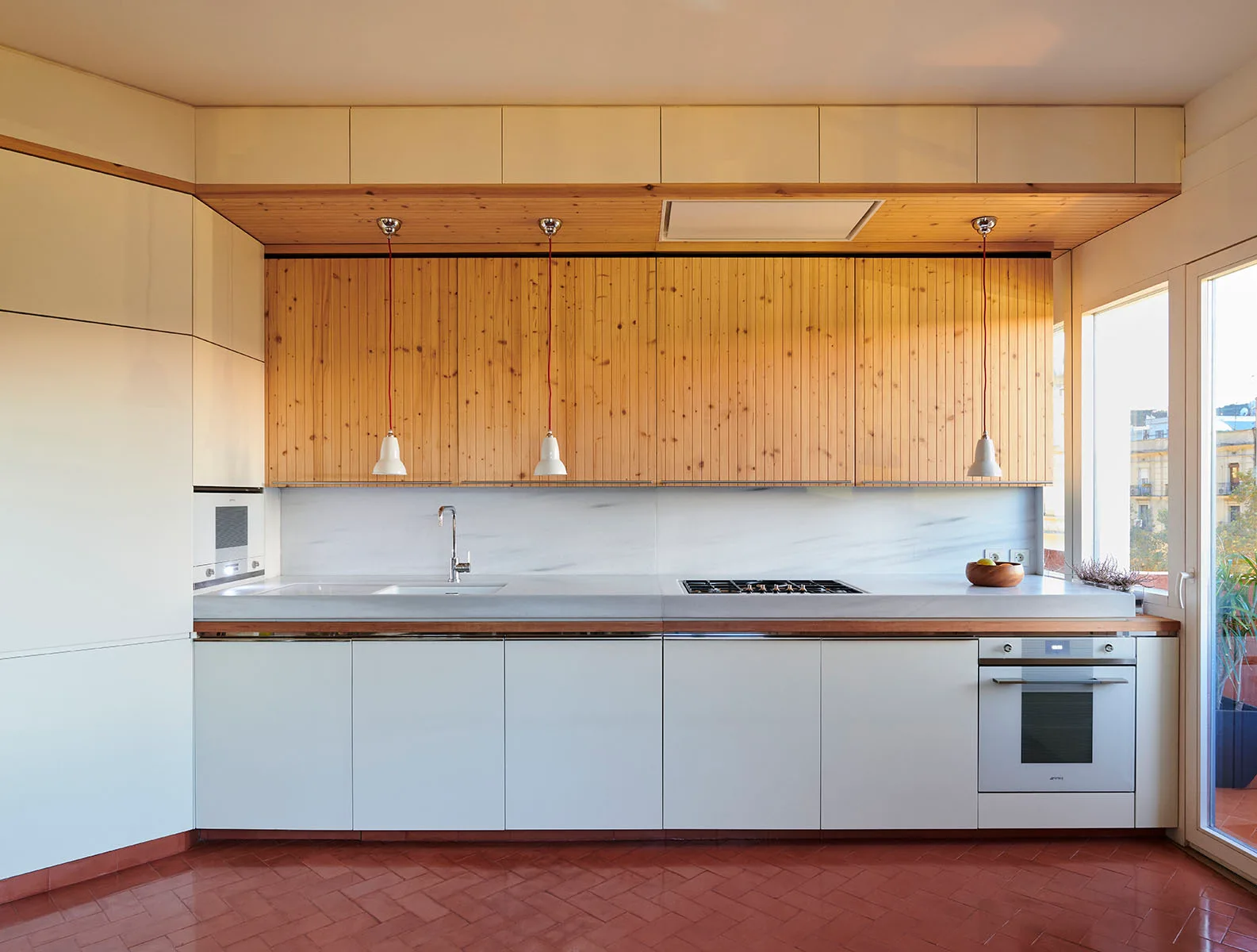A minimalist kitchen features white base cabinets, vertical-slatted wood upper cabinetry, and a red herringbone tile floor.