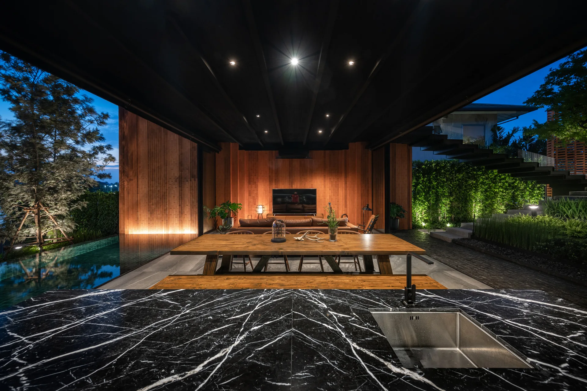 A covered outdoor patio features a black marble kitchen island in the foreground, a wood dining table, and a pool lit at dusk.