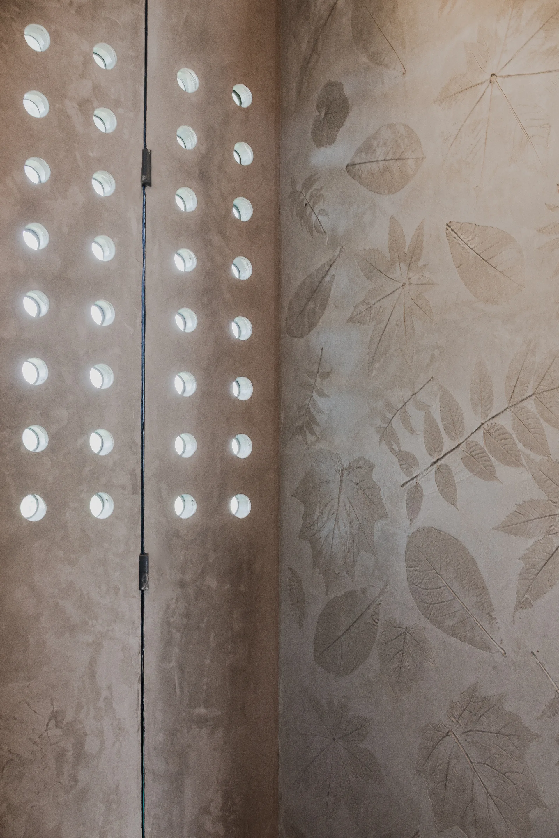 A beige plaster wall features embossed botanical leaf patterns meeting a partition panel with rows of circular glass block inserts.