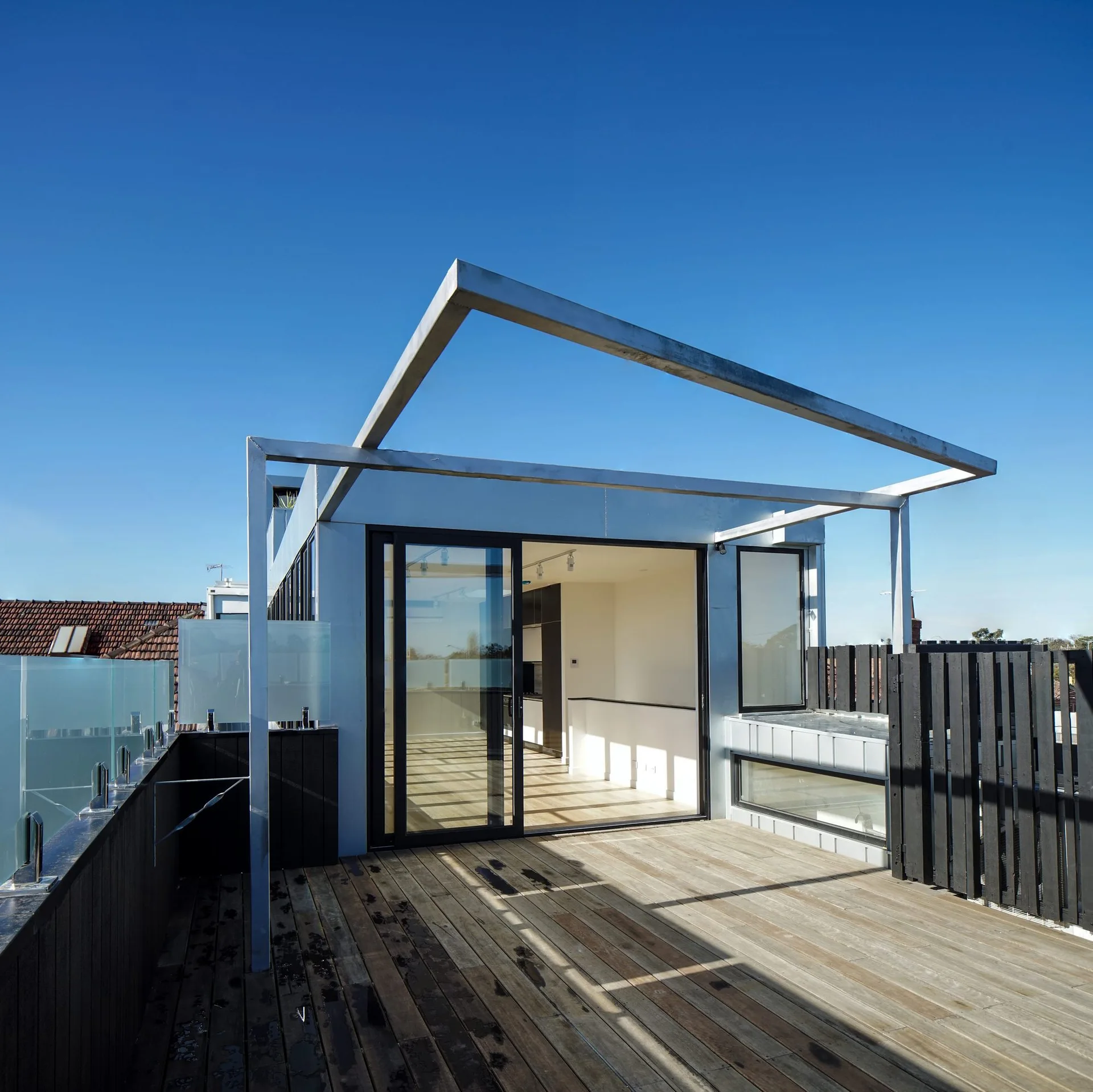 A modern wooden rooftop deck features a minimalist metal frame pergola, a black wood fence, and glass sliding doors leading indoors.