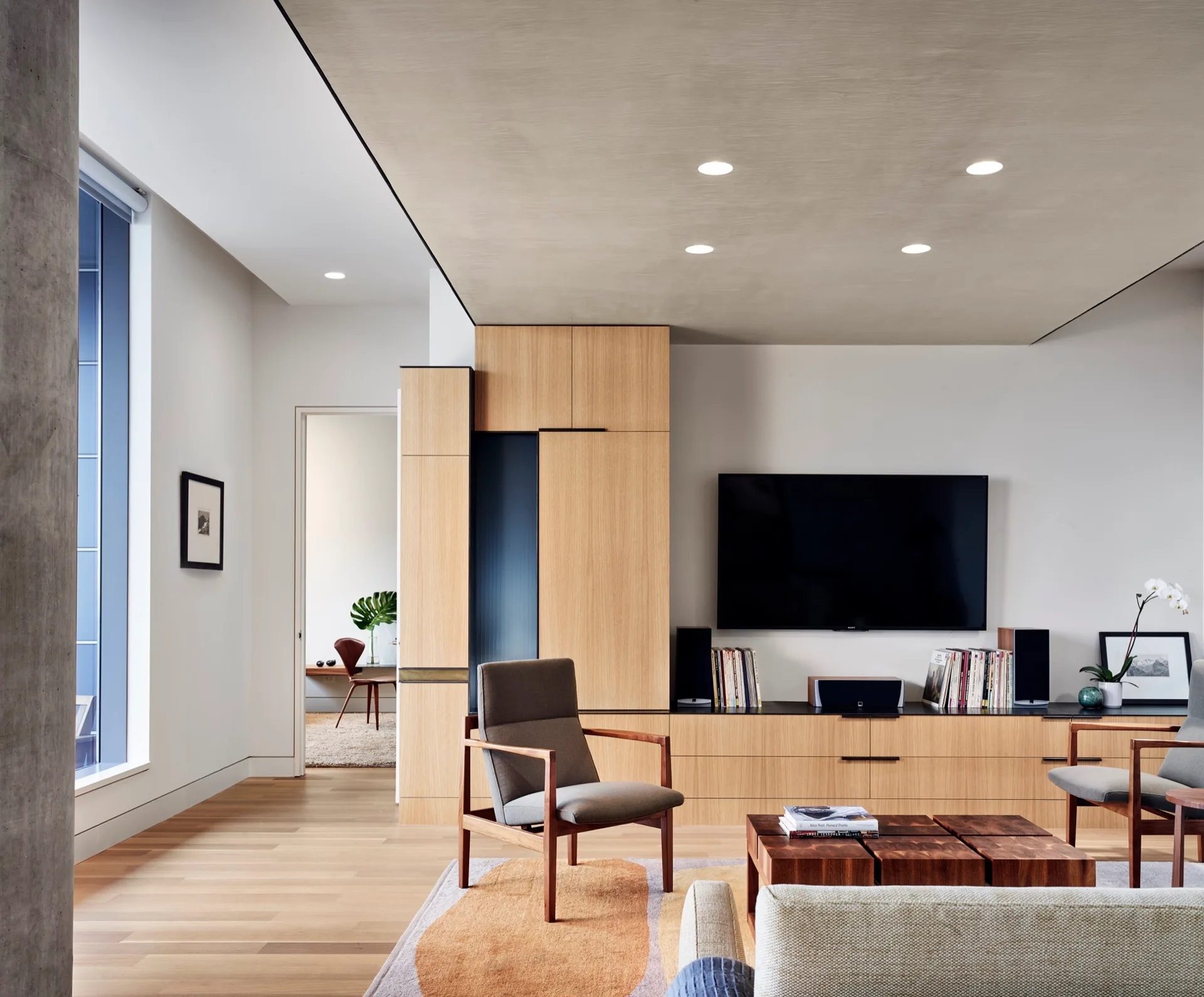 A modern living room features oak built-in cabinetry, grey wood-framed armchairs, a low block coffee table, and an abstract patterned rug.