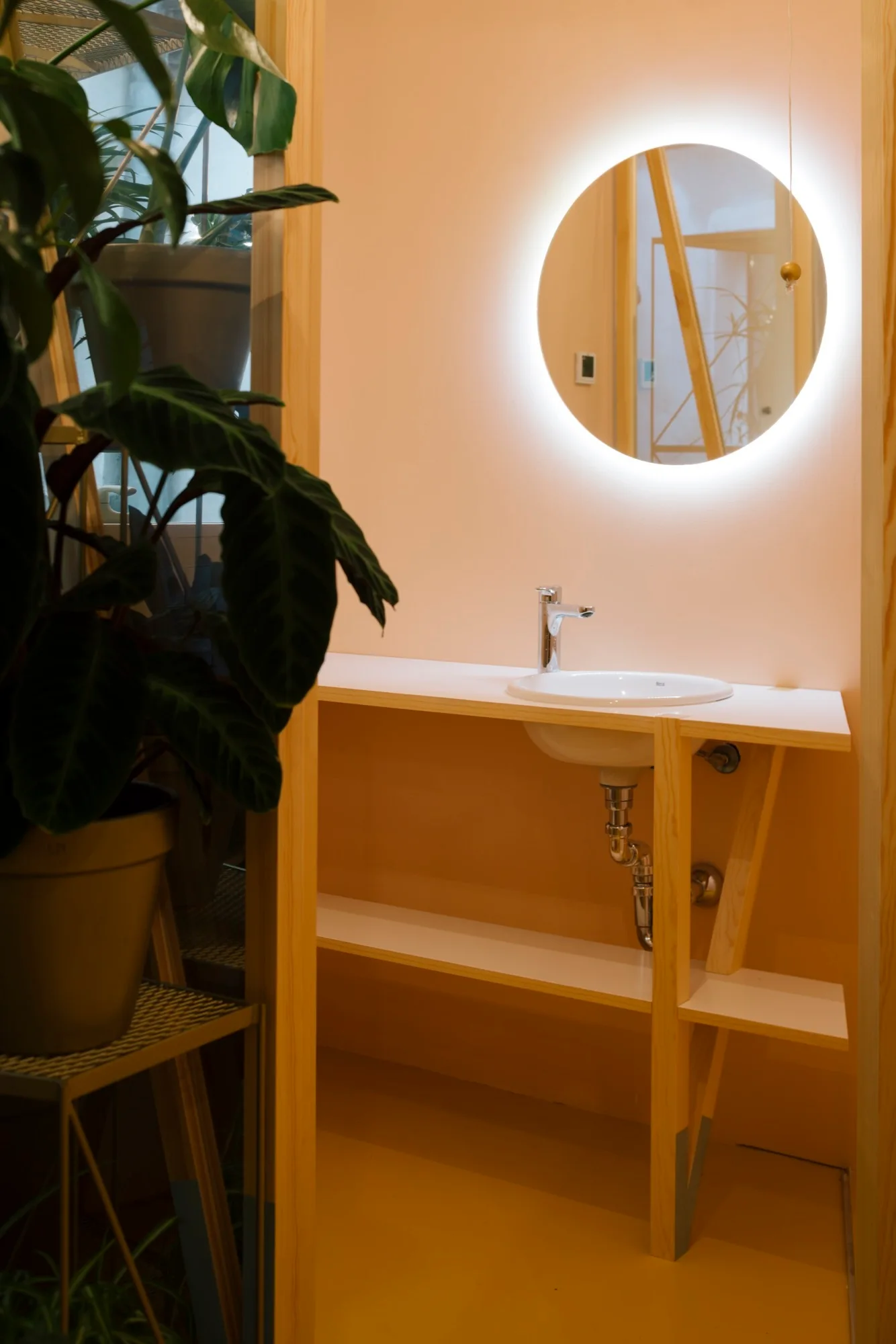 A small white ceramic sink sits in a light wood vanity beneath a glowing, backlit circular mirror against a peach-colored wall.