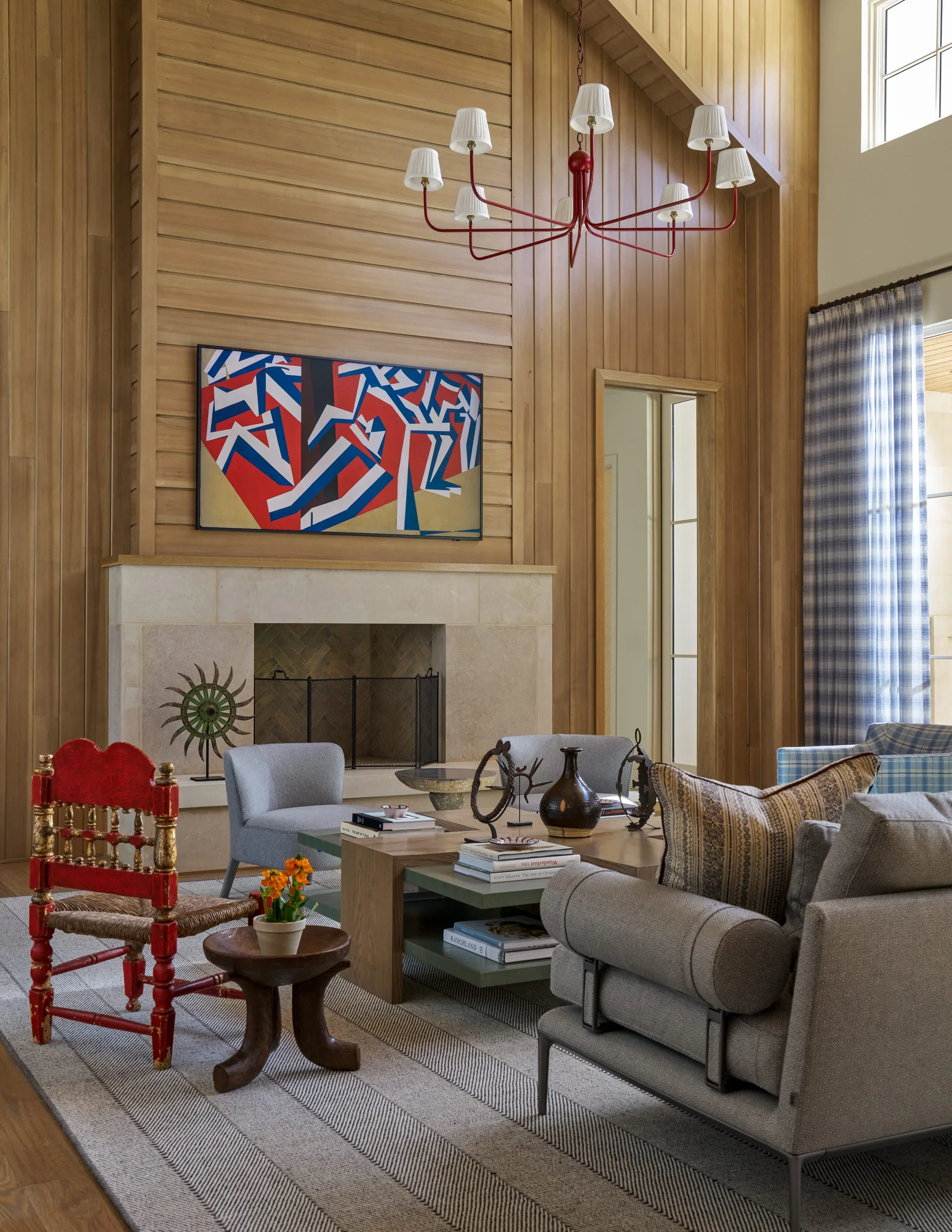 A rustic living room features vertical wood paneling, a stone fireplace, gray armchairs, and a prominent red chandelier hanging from the vaulted ceiling.