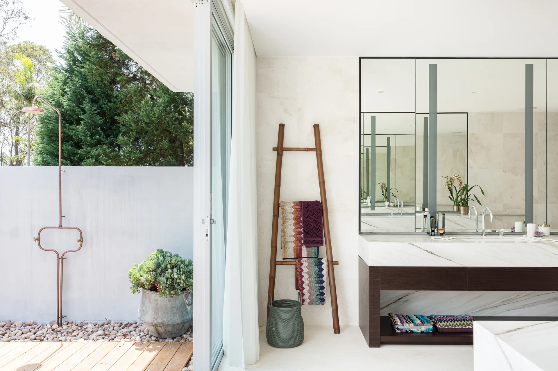 A modern bathroom with a marble vanity and wooden towel ladder next to an open sliding door leading to an outdoor shower area.