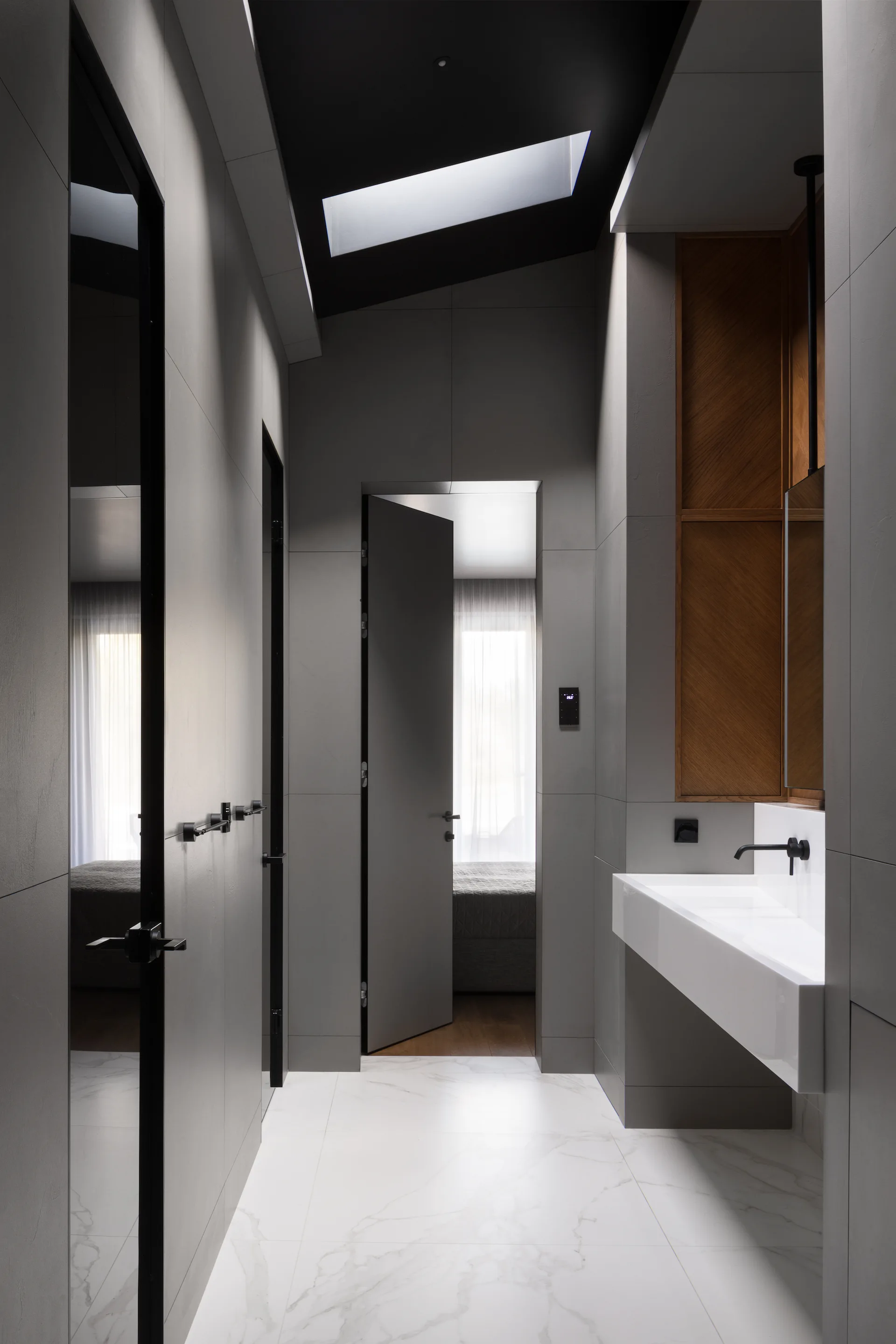 A minimalist grey bathroom featuring a white wall-mounted sink, a skylight, marble floor tiles, and an open doorway leading to a bedroom.