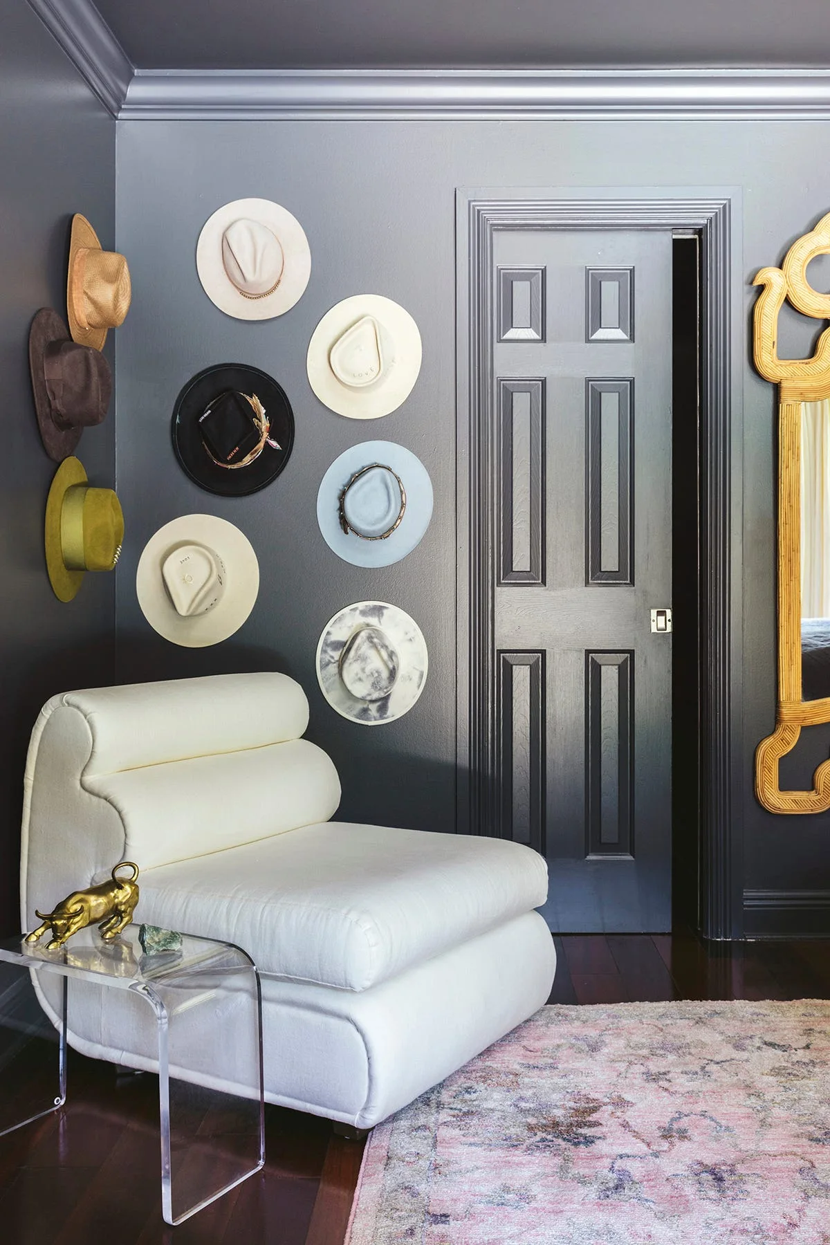 A modern white armchair sits in a dark charcoal bedroom corner next to a gallery wall display of hats and an acrylic side table.