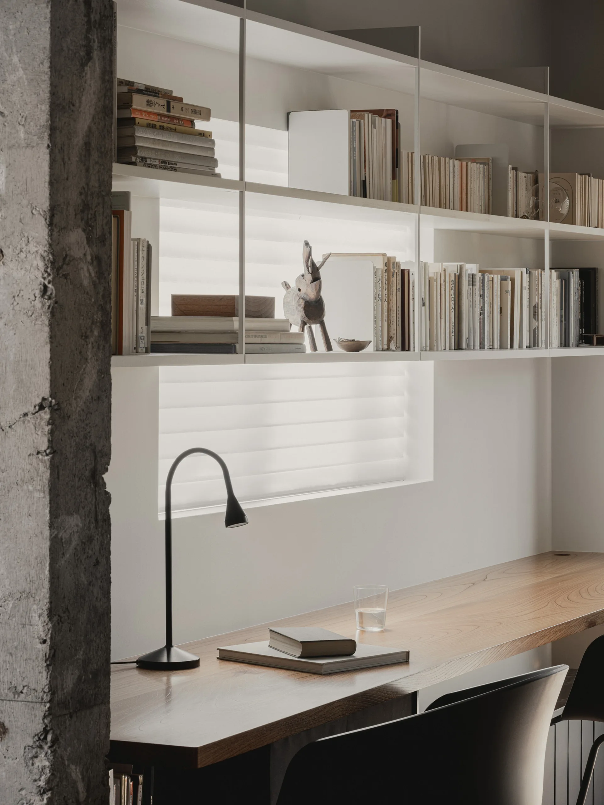 A minimalist home office features a natural wood desk, a slim black desk lamp, and white open shelving mounted across a window.