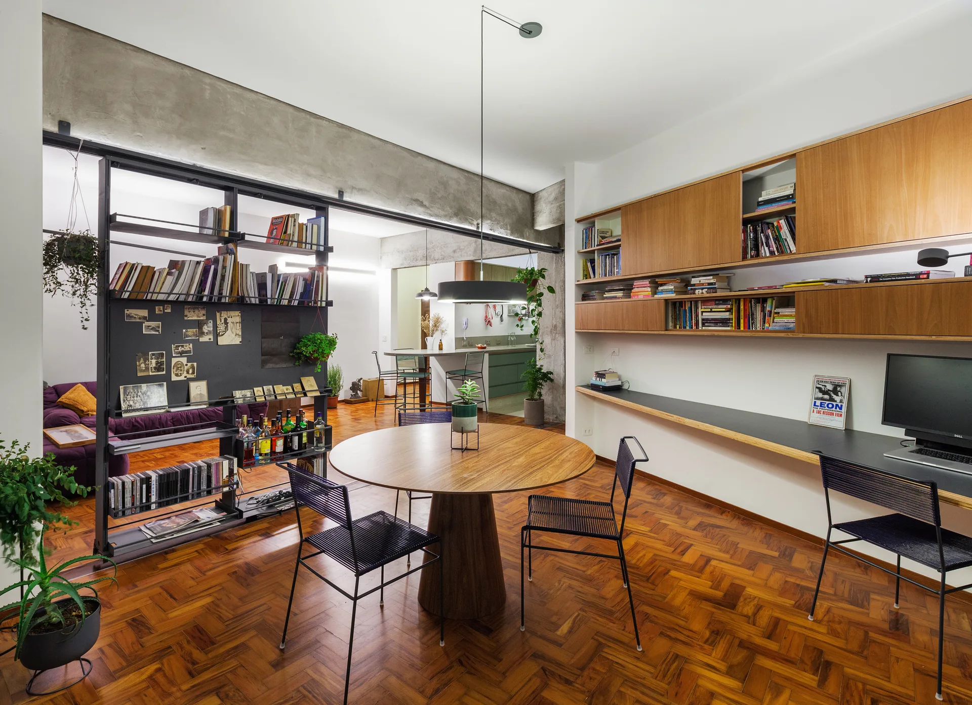 An open-plan dining room features a round wooden table, black wire chairs, a built-in desk, and an open metal room divider over parquet flooring.