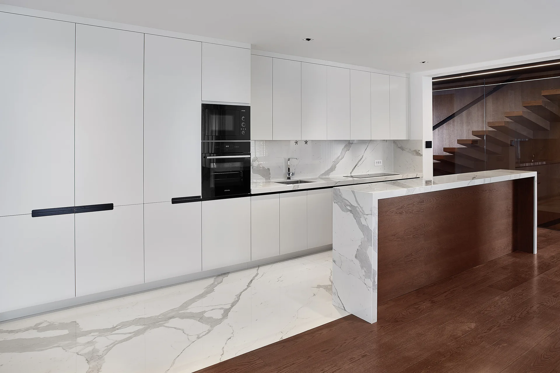 A minimalist kitchen features full-height white cabinets, integrated black appliances, a marble-topped island with a wood front, and veined stone flooring.