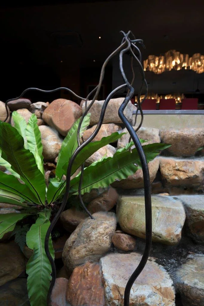 A stacked stone wall features a green fern and a winding dark metal sculpture in the foreground, with warm background lighting.