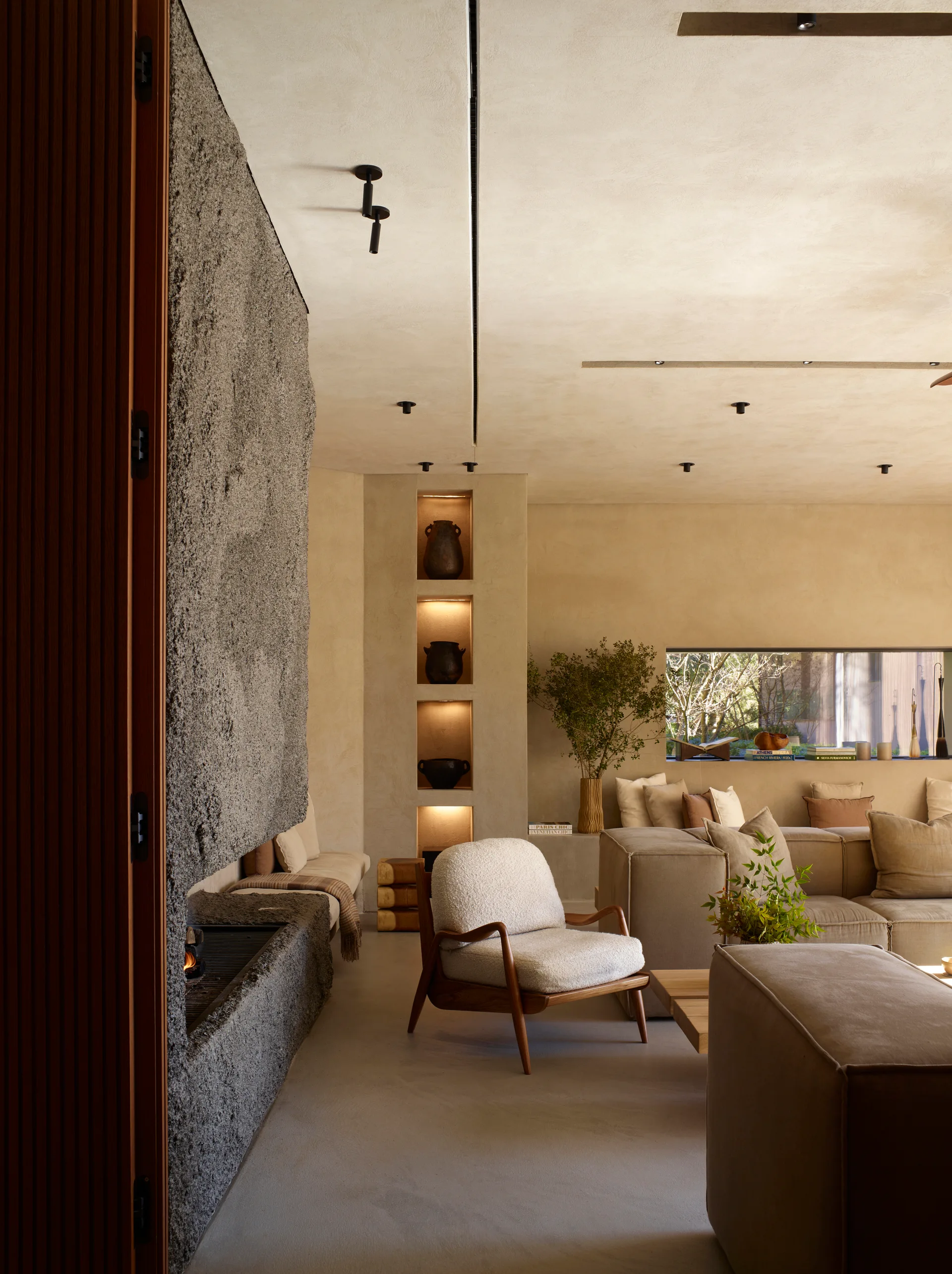 A minimalist living room features a rough-textured stone fireplace, beige plaster walls with lighted display niches, and a modular neutral sofa.