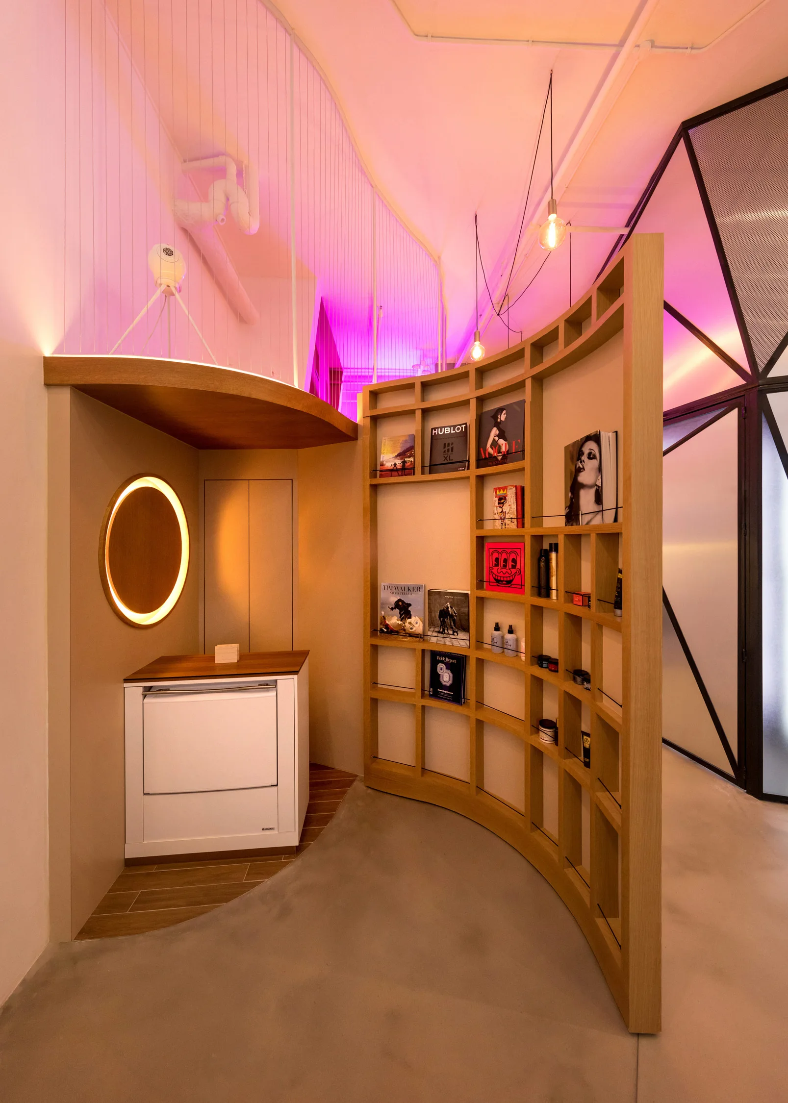 A curved wooden grid shelving unit stands next to a small beige niche featuring a circular backlit mirror and white cabinet.