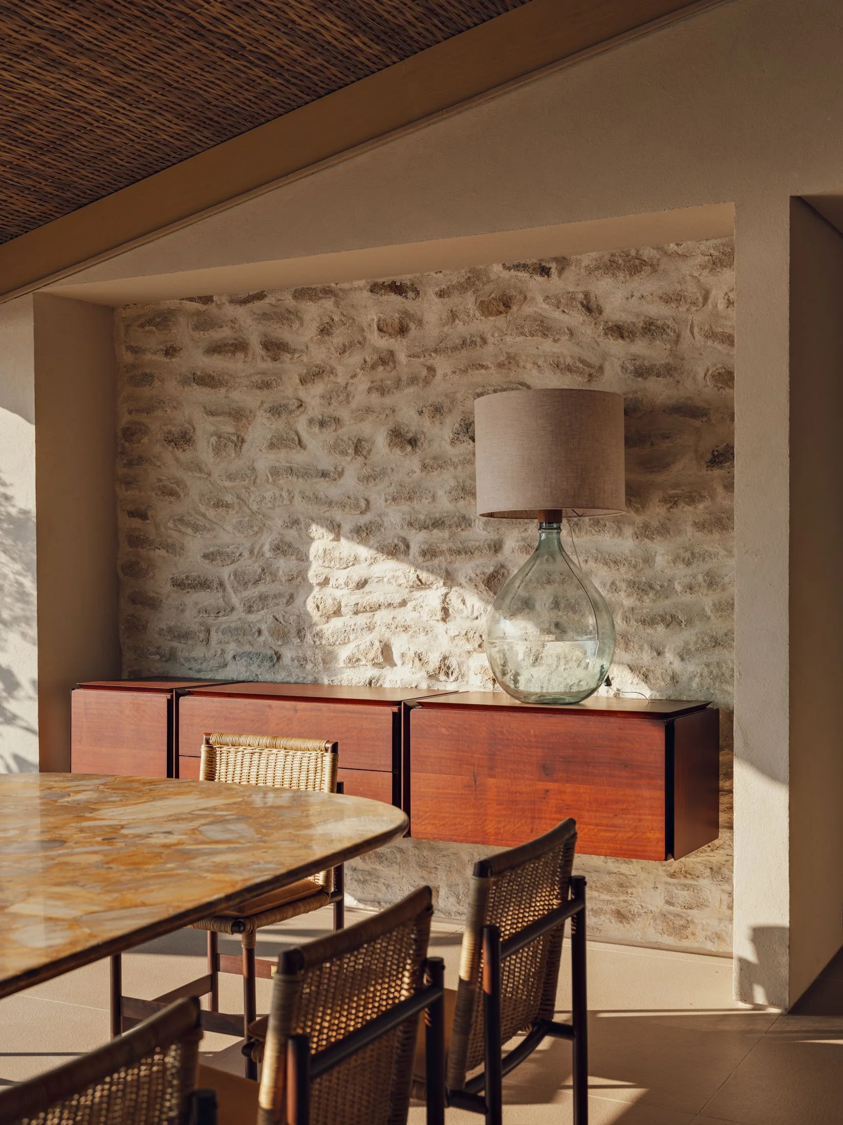 A dining area features a textured stone wall, a floating wooden sideboard topped with a glass lamp, and a marble-topped table with woven chairs.