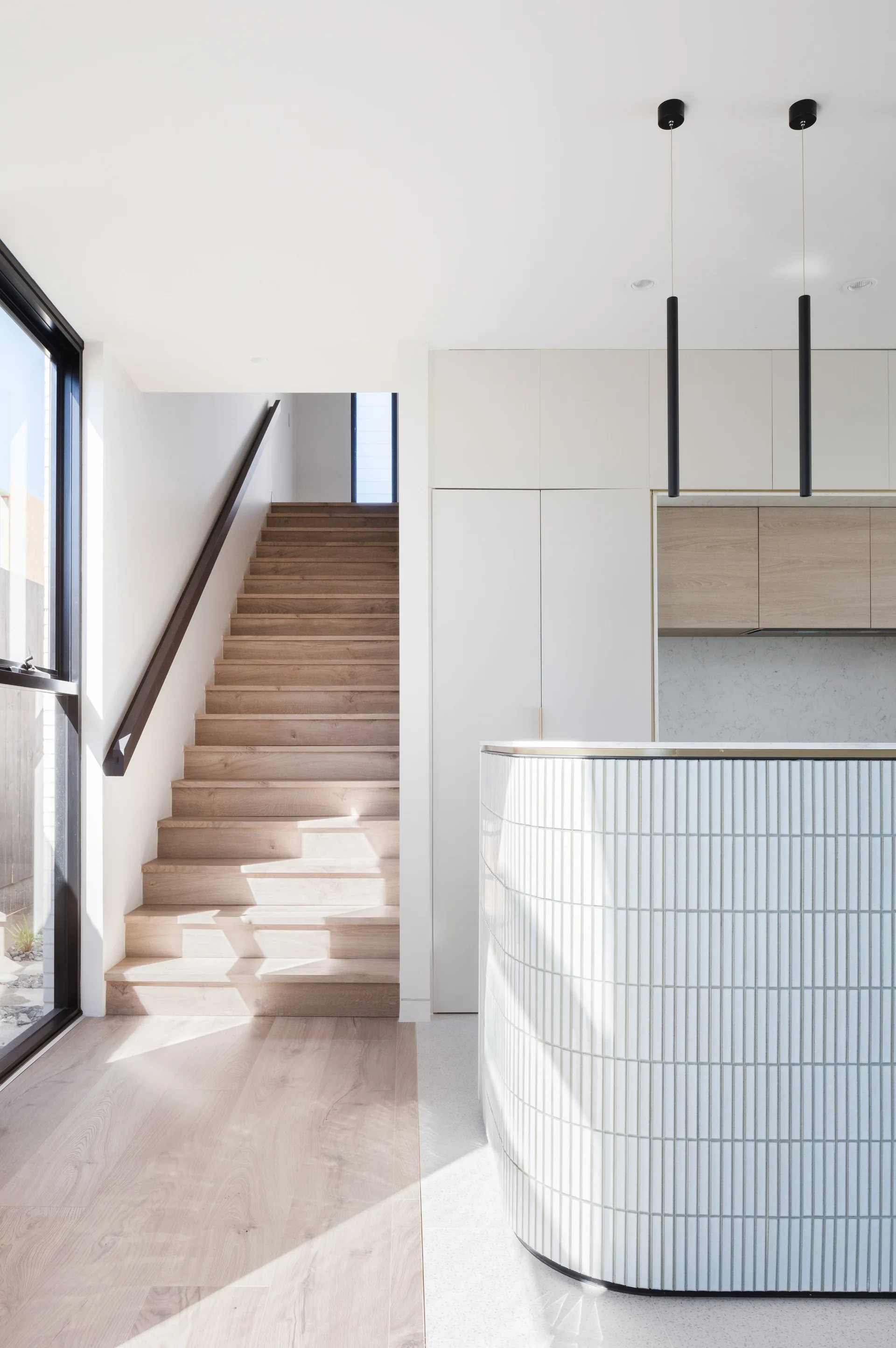 A light wood staircase ascends next to a curved kitchen island clad in vertical white tiles under black pendant lights.