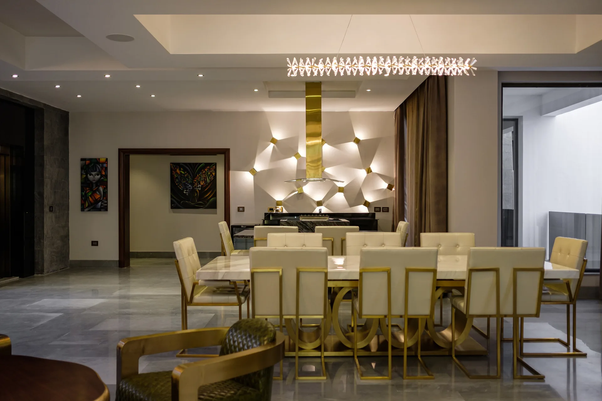 A dining room features a marble table surrounded by cream chairs with gold frames, set before an illuminated geometric wall with a gold range hood.