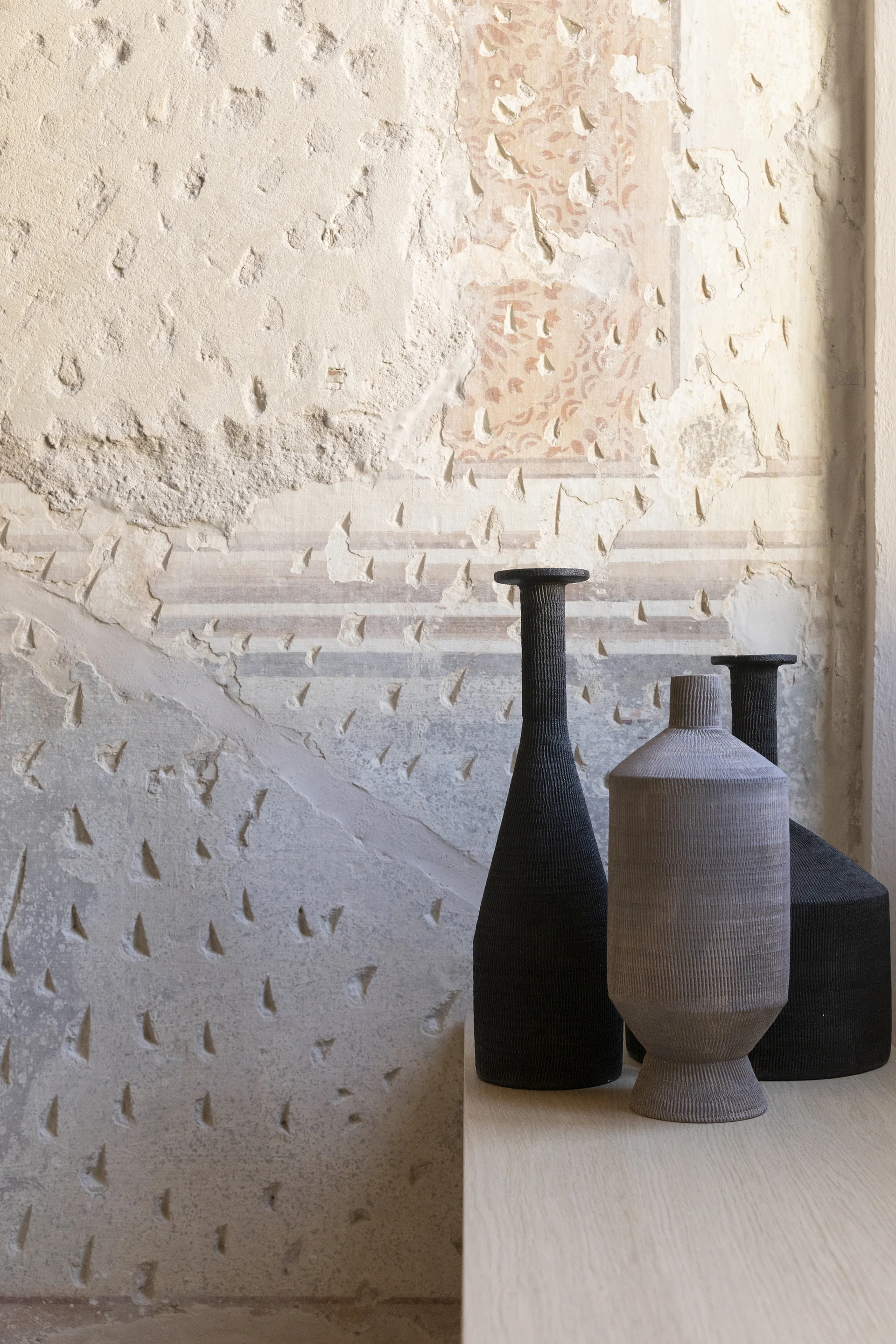 Three ribbed ceramic vases in black and grey stand on a light wood shelf in front of a heavily distressed beige plaster wall.