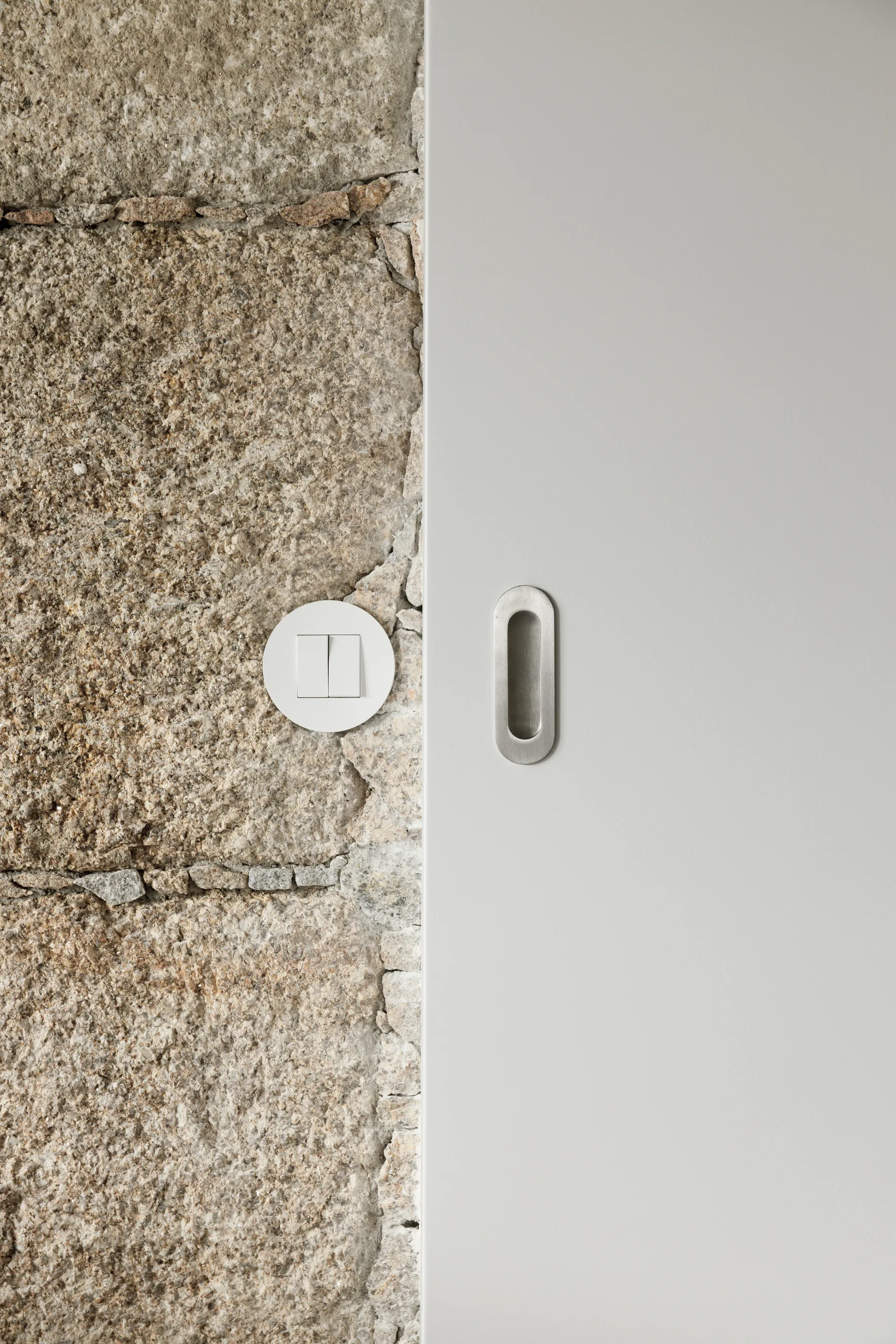 A close-up of a rugged stone wall featuring a round white switch transitioning to a smooth grey panel with a recessed metal handle.