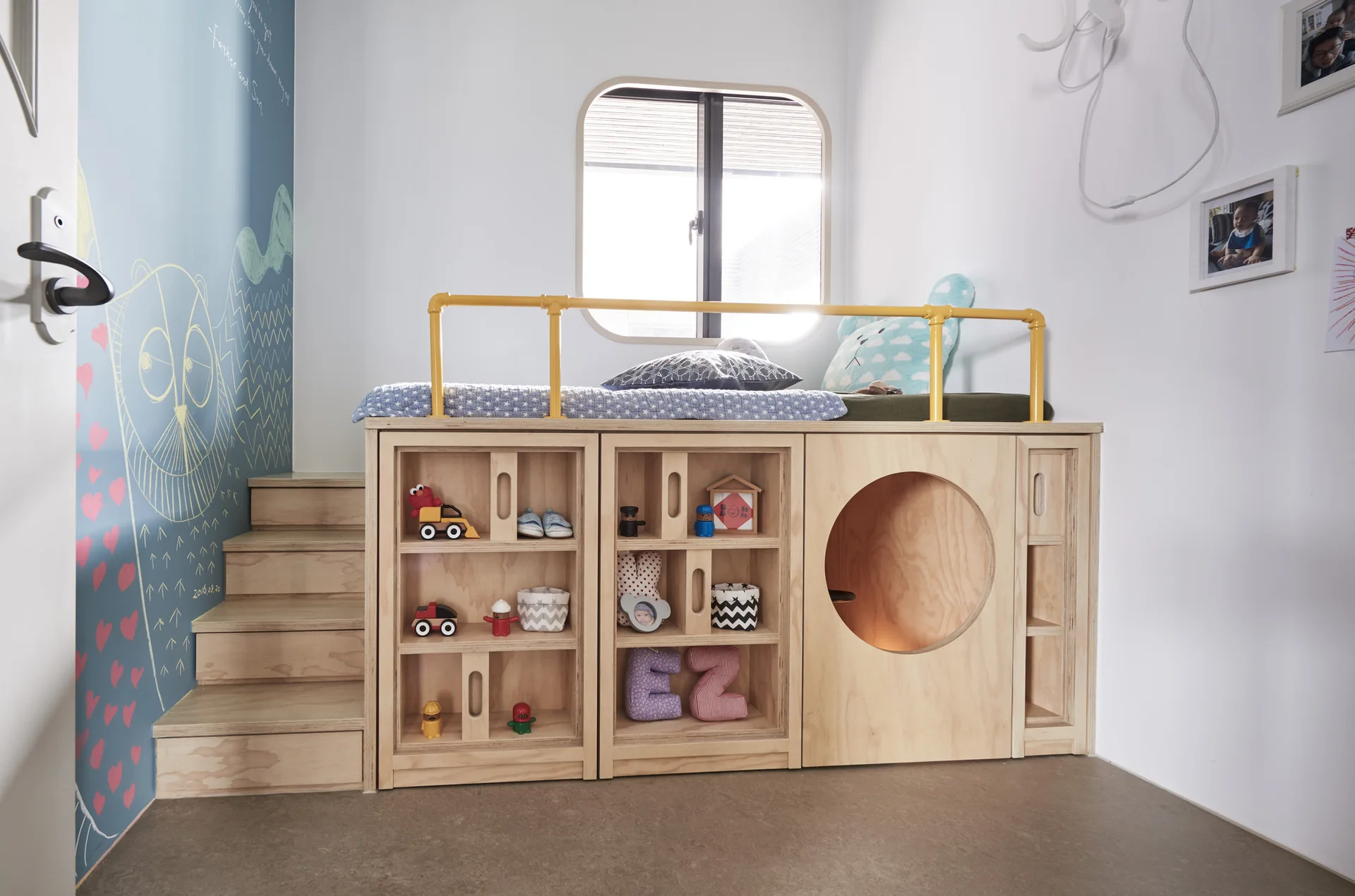 A custom plywood loft bed features integrated toy shelves, a circular playroom cutout, wooden steps, and a bright yellow safety railing.