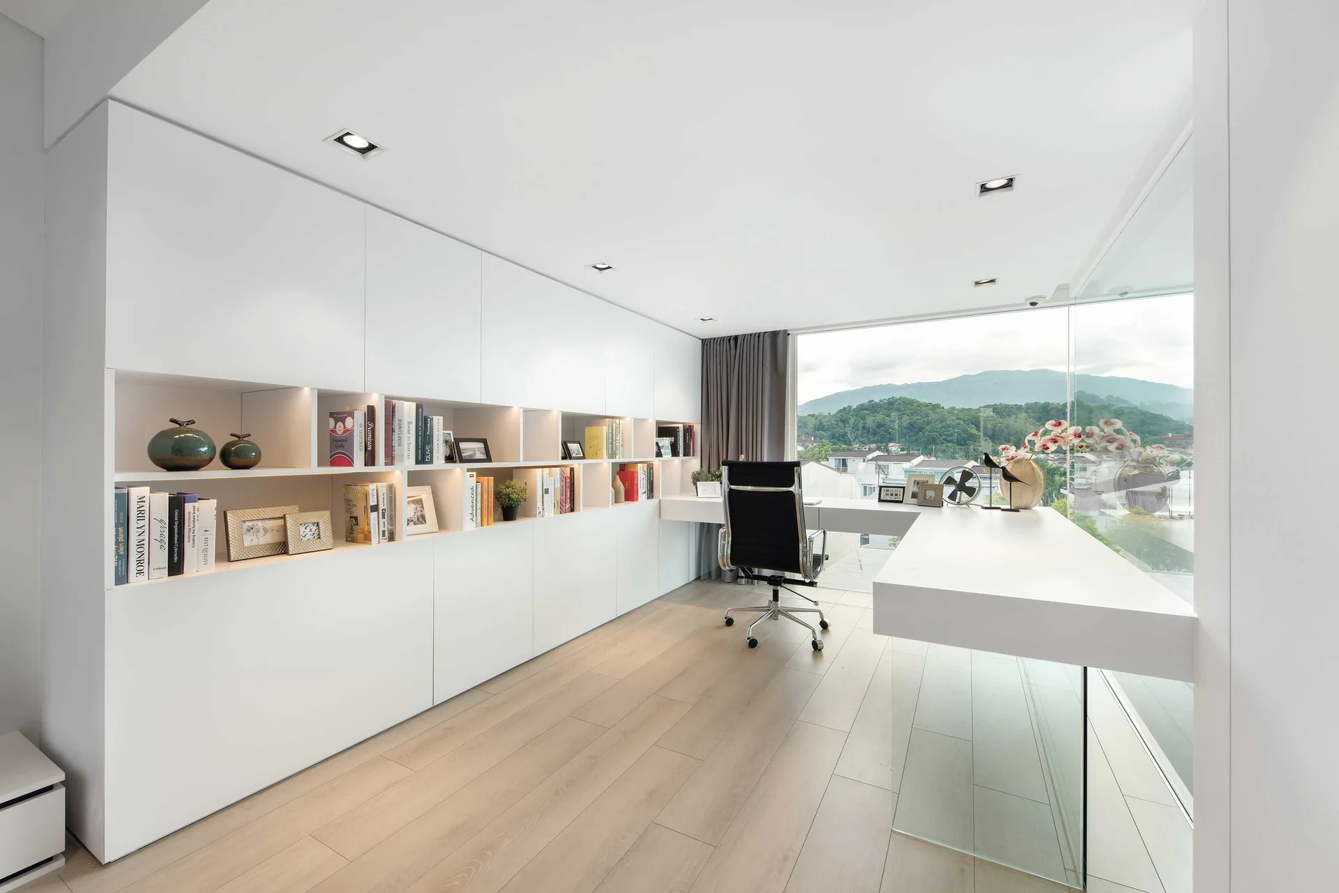 A minimalist home office featuring a long white desk with glass support, integrated floor-to-ceiling cabinets with books, and a large window.
