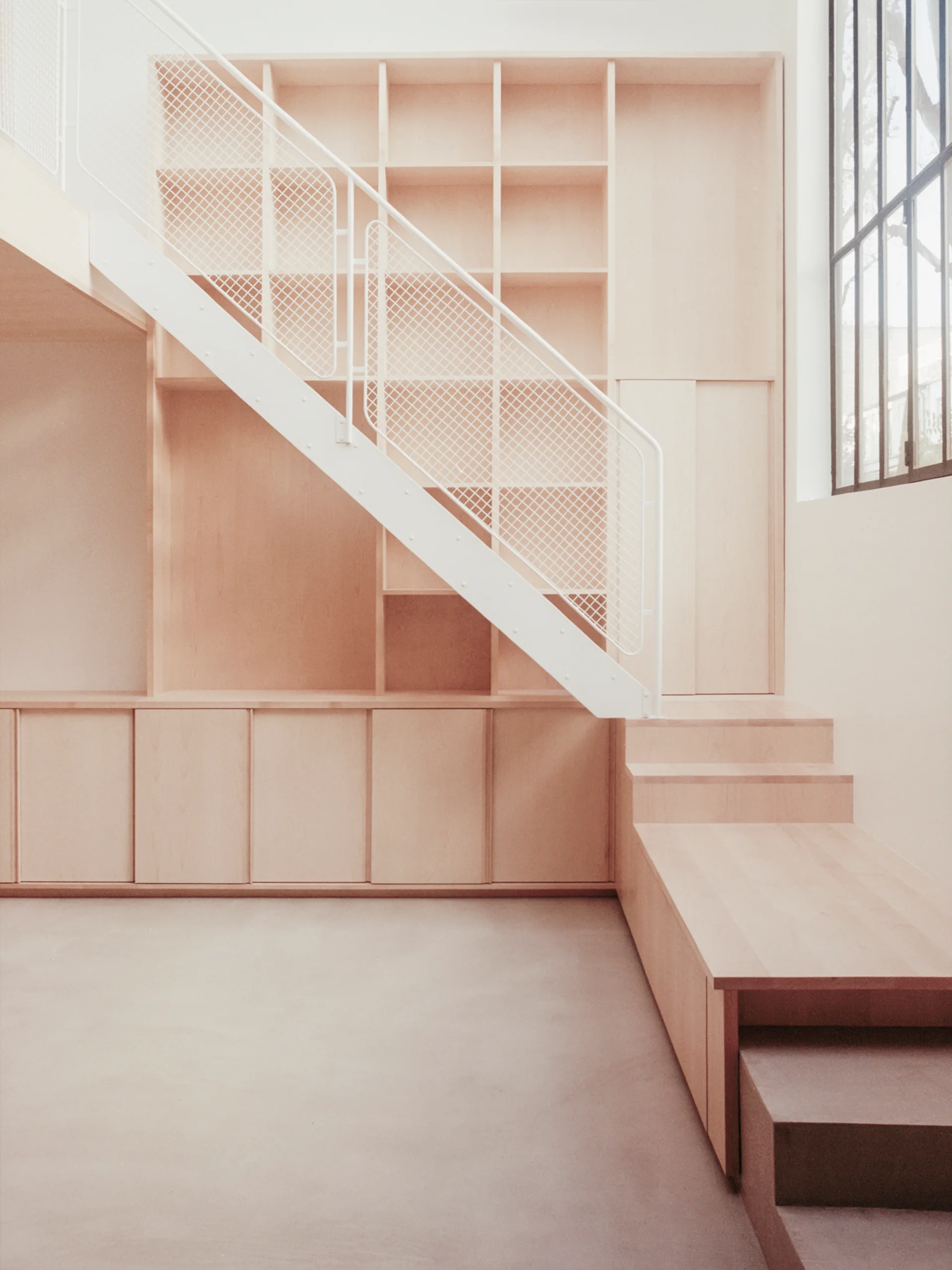 A minimalist light wood staircase featuring a white metal mesh railing integrates seamlessly with matching built-in shelving and low cabinetry.