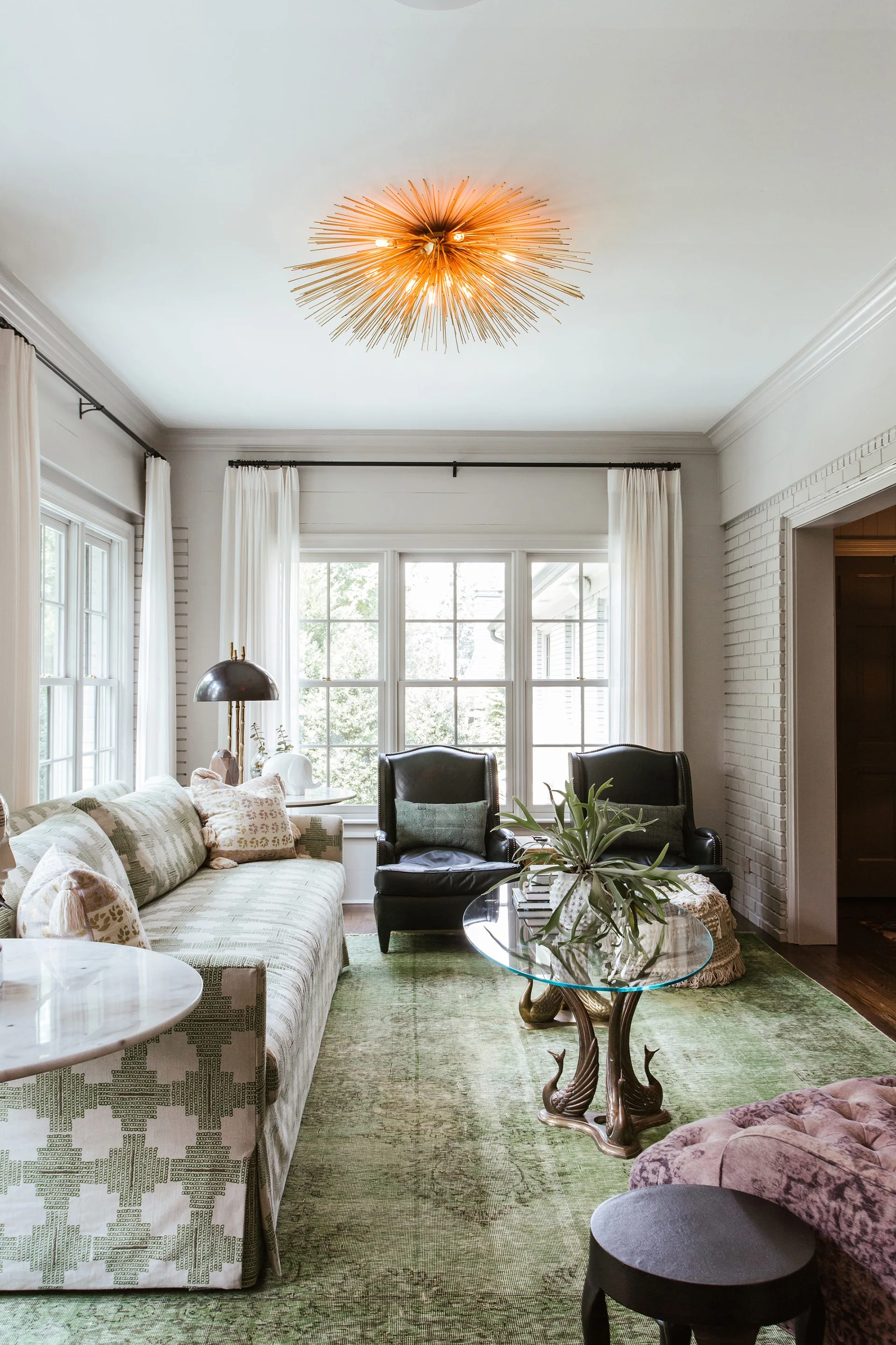 An eclectic living room featuring a patterned sofa, dark leather armchairs, a glass coffee table, and a green distressed area rug.