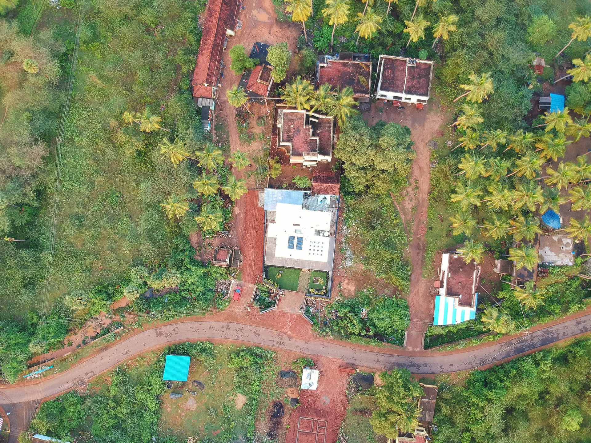 An aerial top-down view of a rural residential property with multiple buildings, dirt pathways, green vegetation, and tropical palm trees.