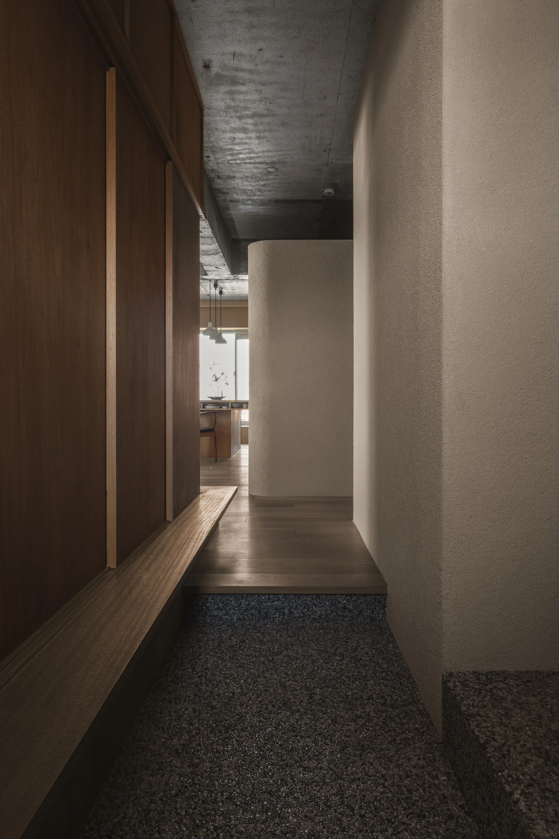 A minimalist hallway features a pebbled entry transition, integrated wood closets with a floating bench, and textured plaster walls.