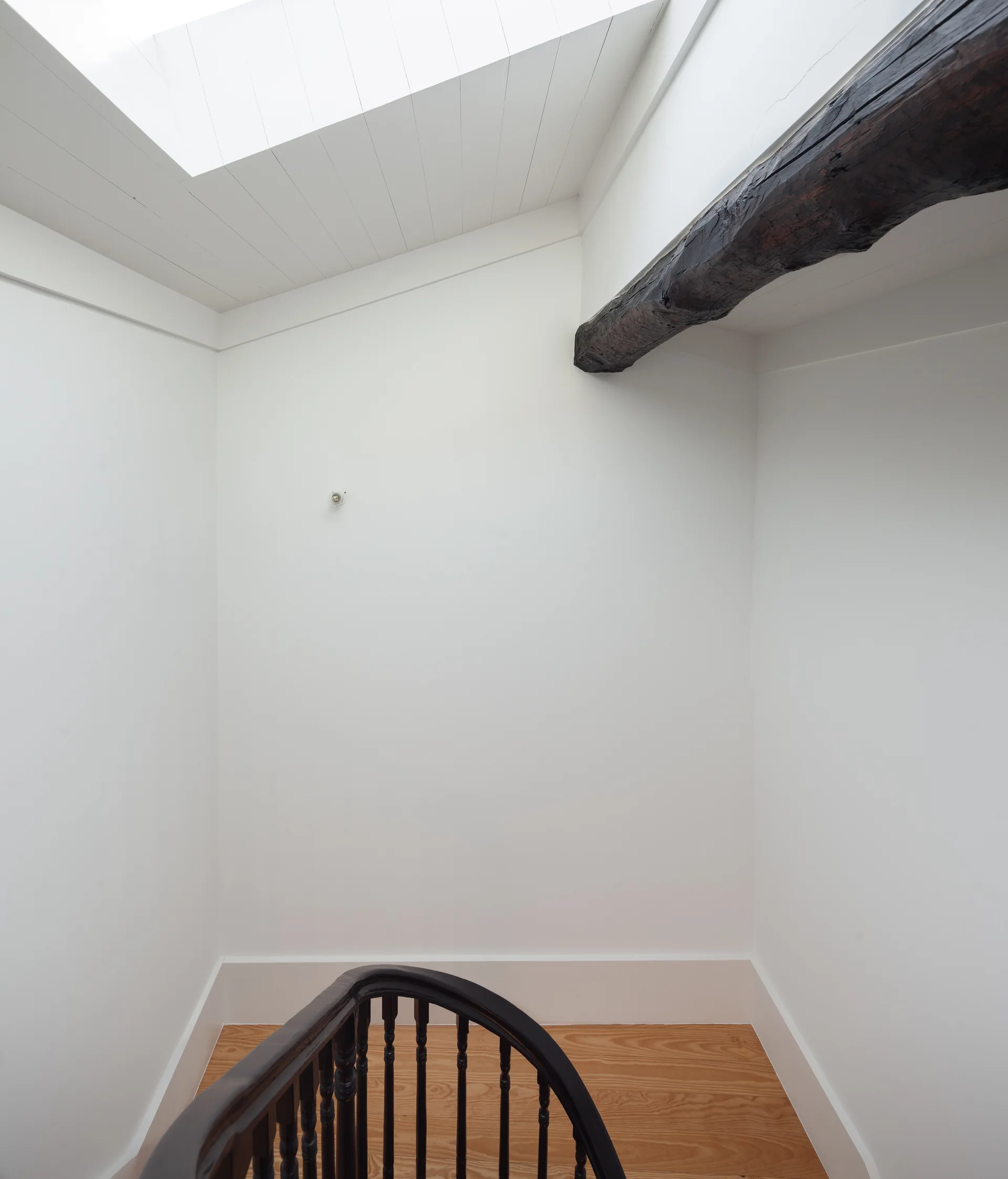 A white staircase landing features a curved dark wood railing, light hardwood flooring, a skylight, and a rustic dark timber beam overhead.