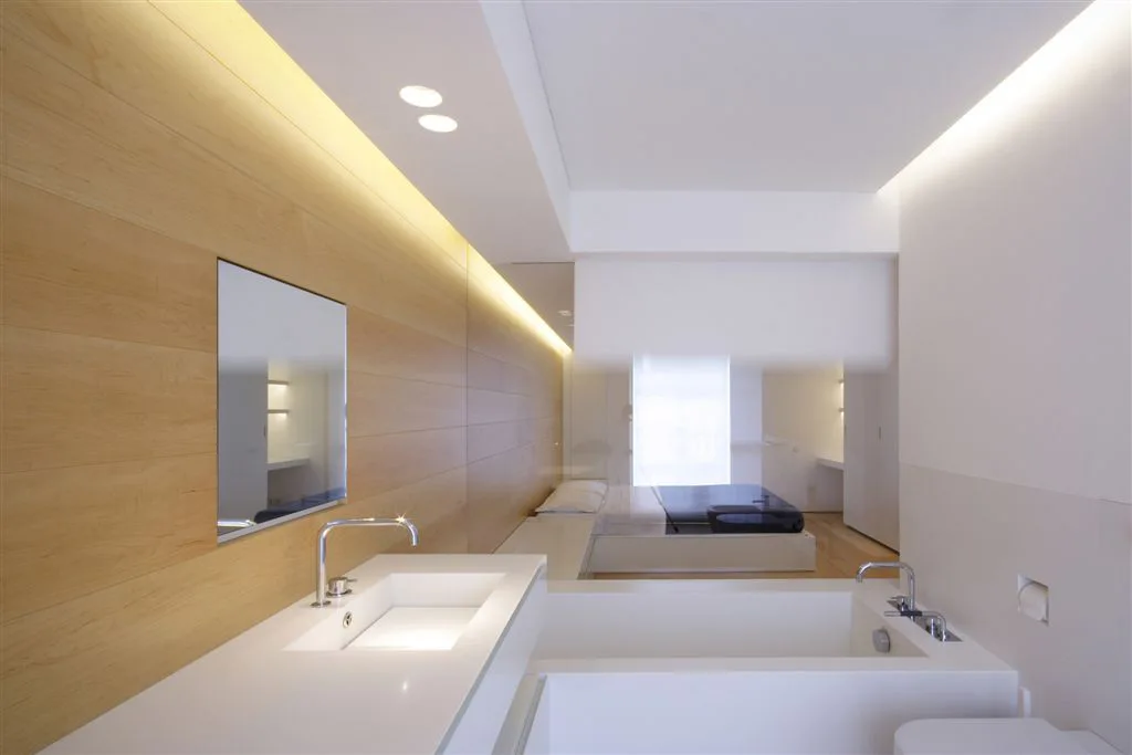 A minimalist white bathroom featuring a light wood paneled wall, integrated vanity, and bathtub separated from a bedroom by a clear glass partition.