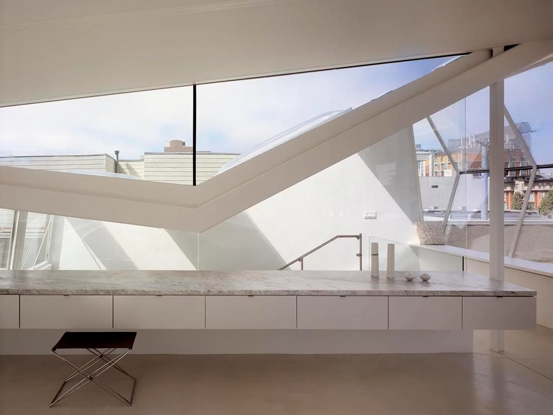 A long built-in white desk with a veined stone top sits beneath a large angular skylight window in a minimalist interior space.