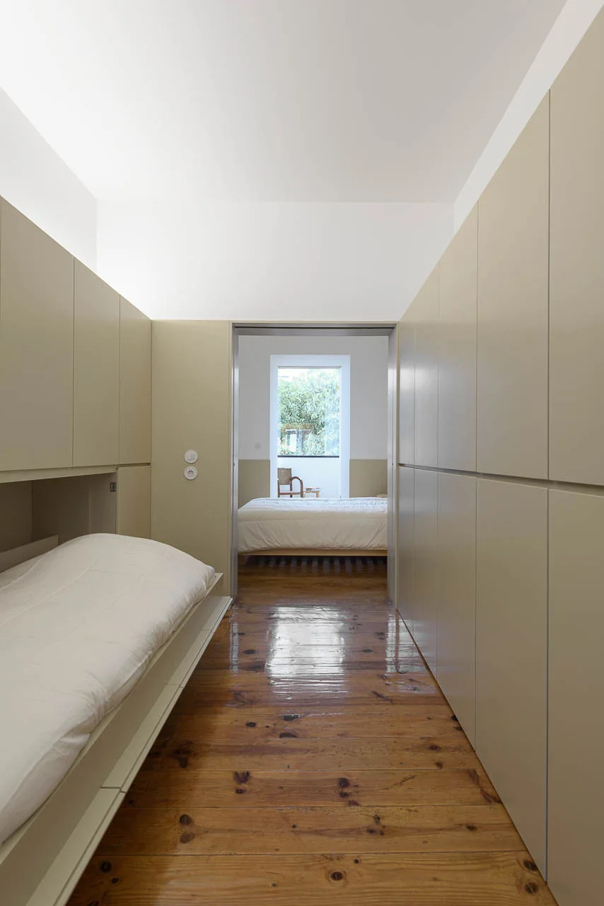 Minimalist bedroom hallway featuring built-in beige cabinetry with an integrated bed on glossy wooden floors leading to a sunlit room.