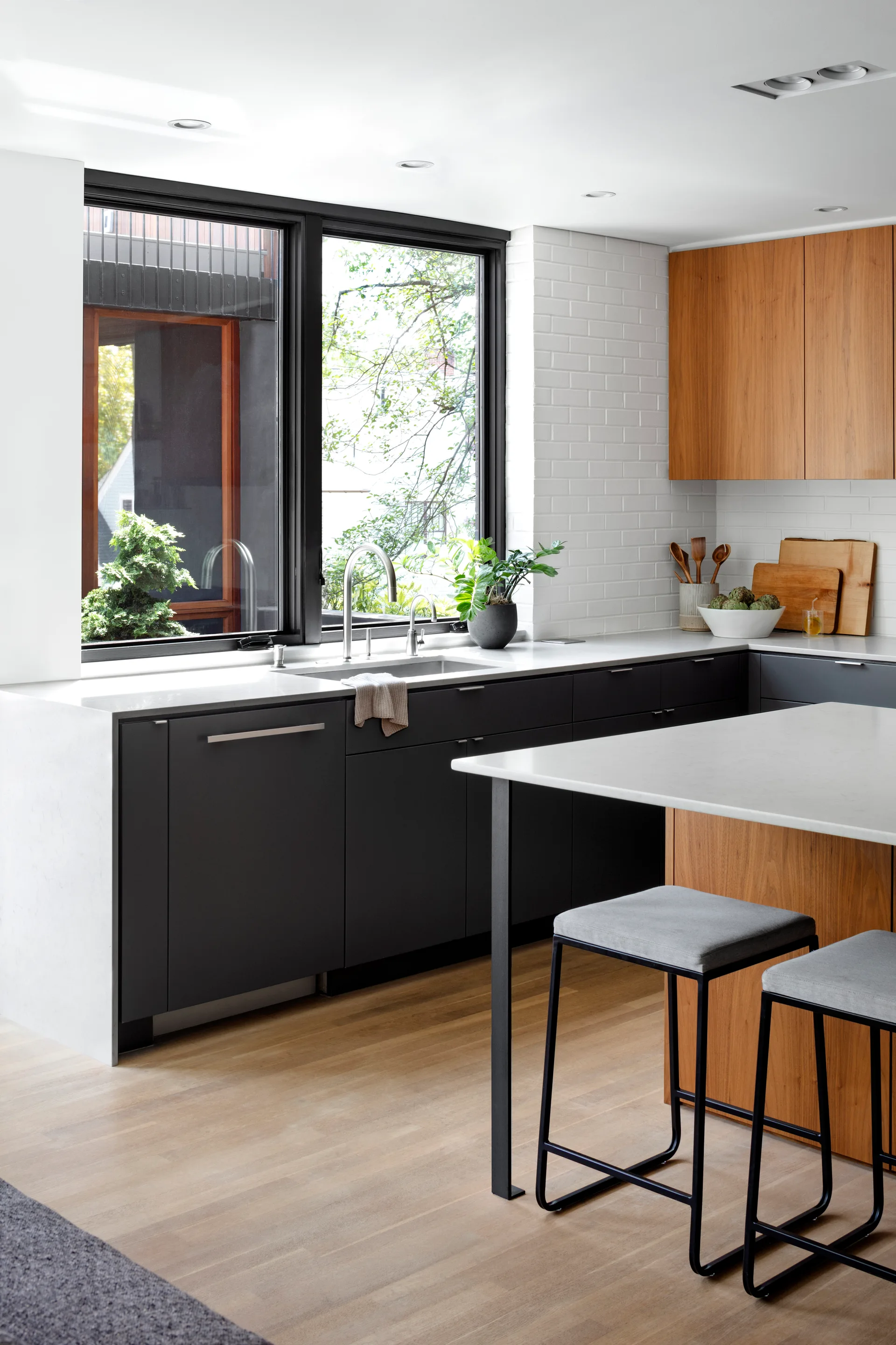 A modern kitchen features dark gray lower cabinets, warm wood upper cabinets, a white brick backsplash, and a central island with bar stools.