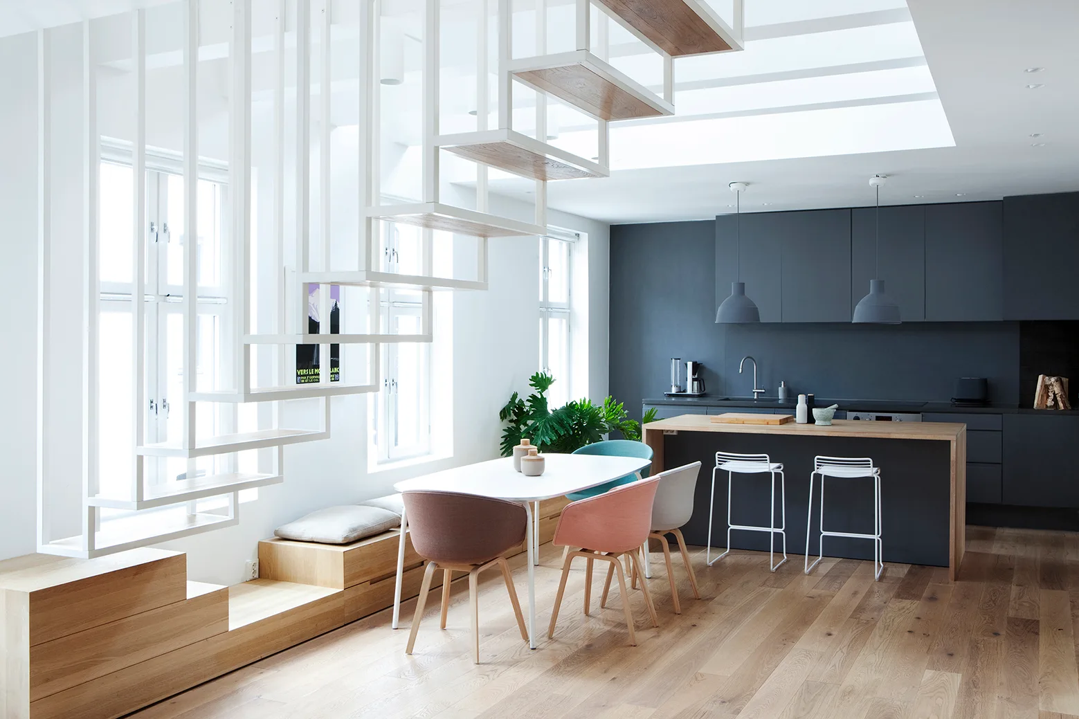 A modern dining area features an open white staircase, a wood platform bench, an oval table with colorful chairs, and a dark grey kitchen island.
