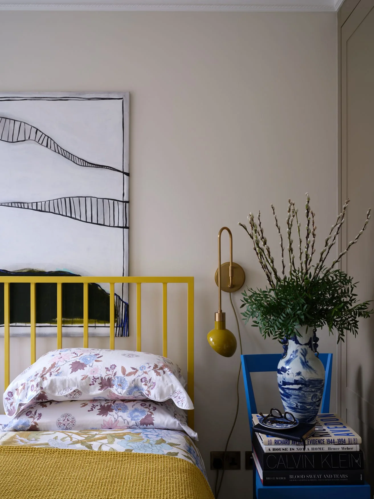 A bedroom corner features a bright yellow metal headboard, floral pillows, a brass wall sconce, and a blue wooden chair used as a nightstand.