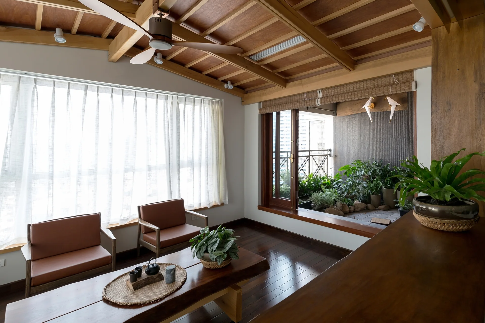 A living room with a vaulted wood ceiling, dark wood floors, two leather armchairs, and a large window looking onto an integrated balcony garden.