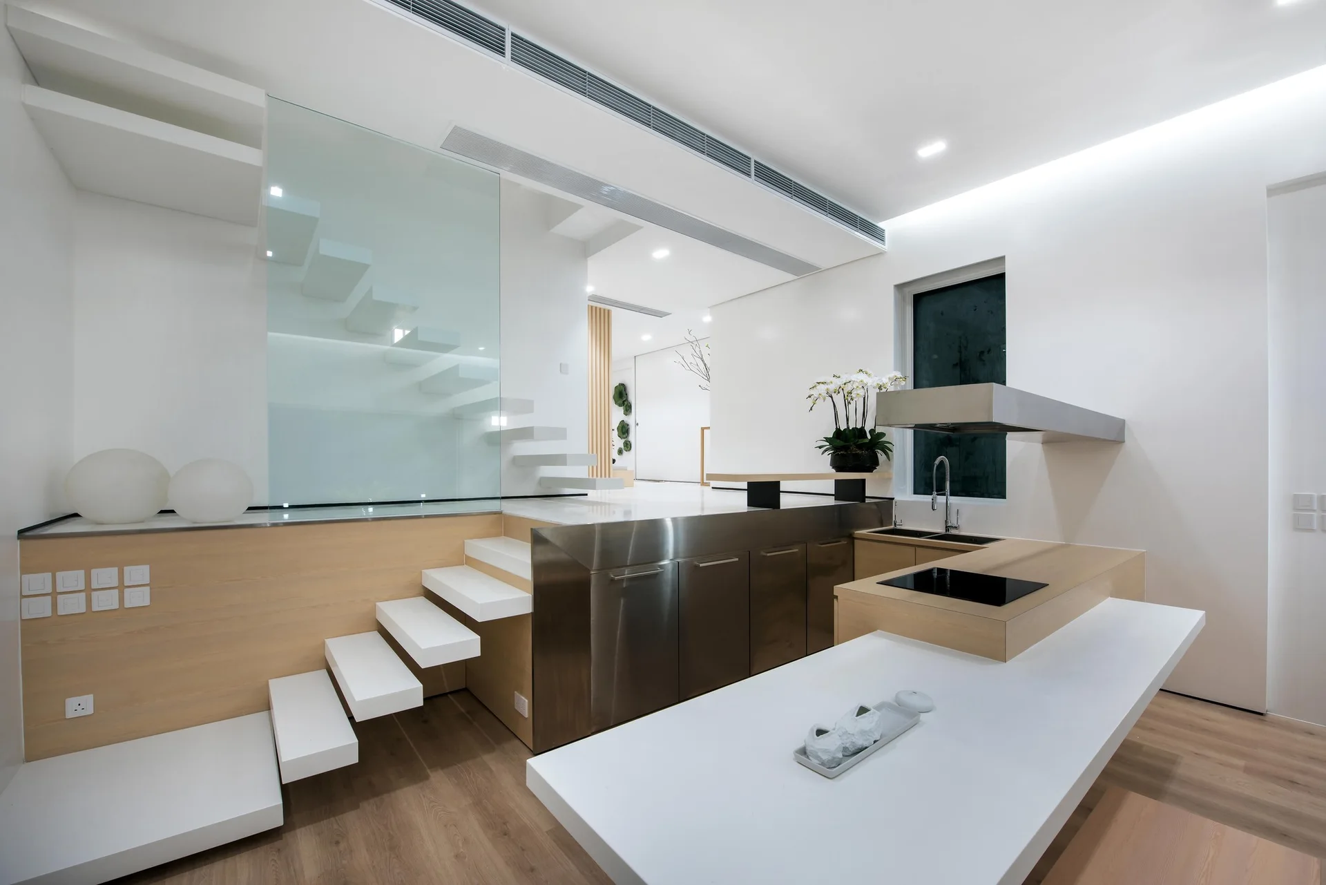 A minimalist kitchen features a white central island, stainless steel cabinetry, and white floating stairs next to a clear glass partition.