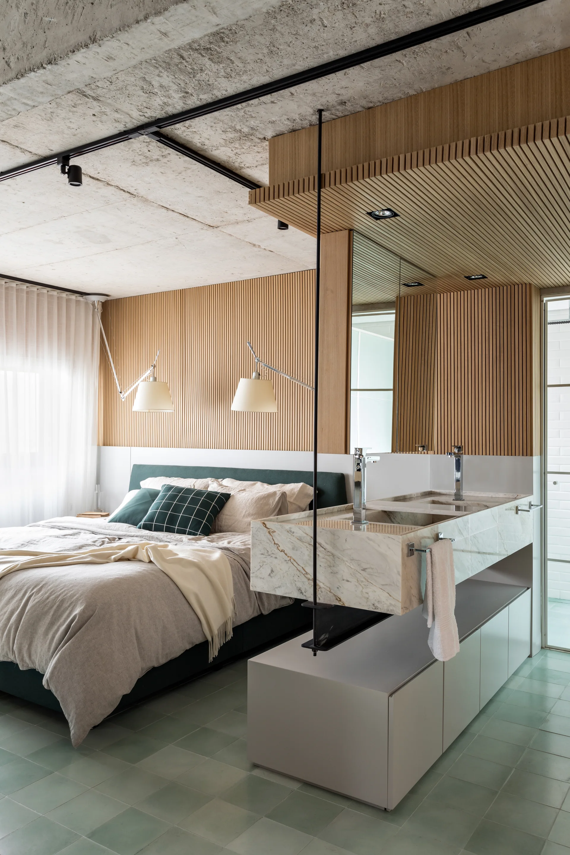 A modern bedroom features a green upholstered bed, raw concrete ceiling, and an integrated marble vanity with a vertical wood slatted accent wall.