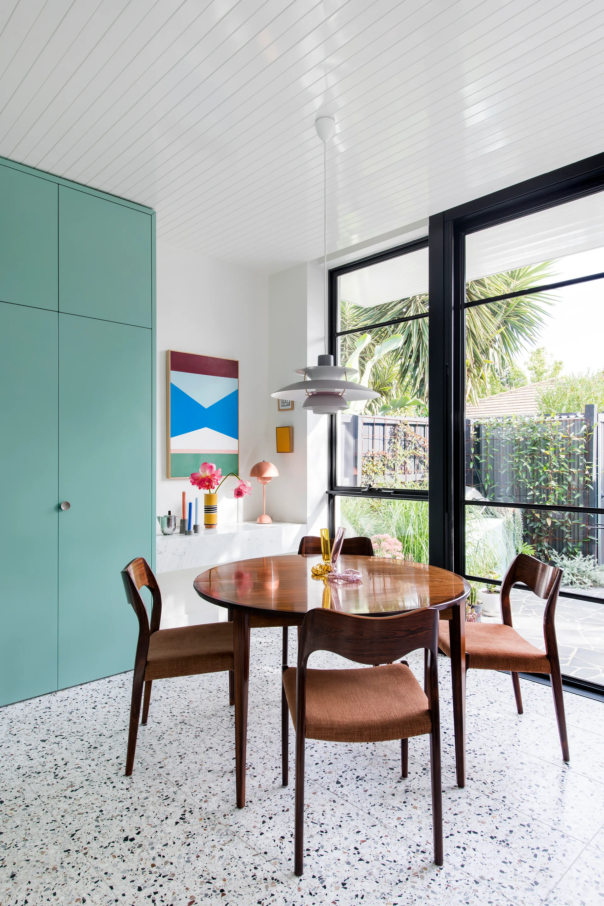 A dining area with a round wooden table, four mid-century modern chairs, large mint green cabinetry, and terrazzo tile floors.