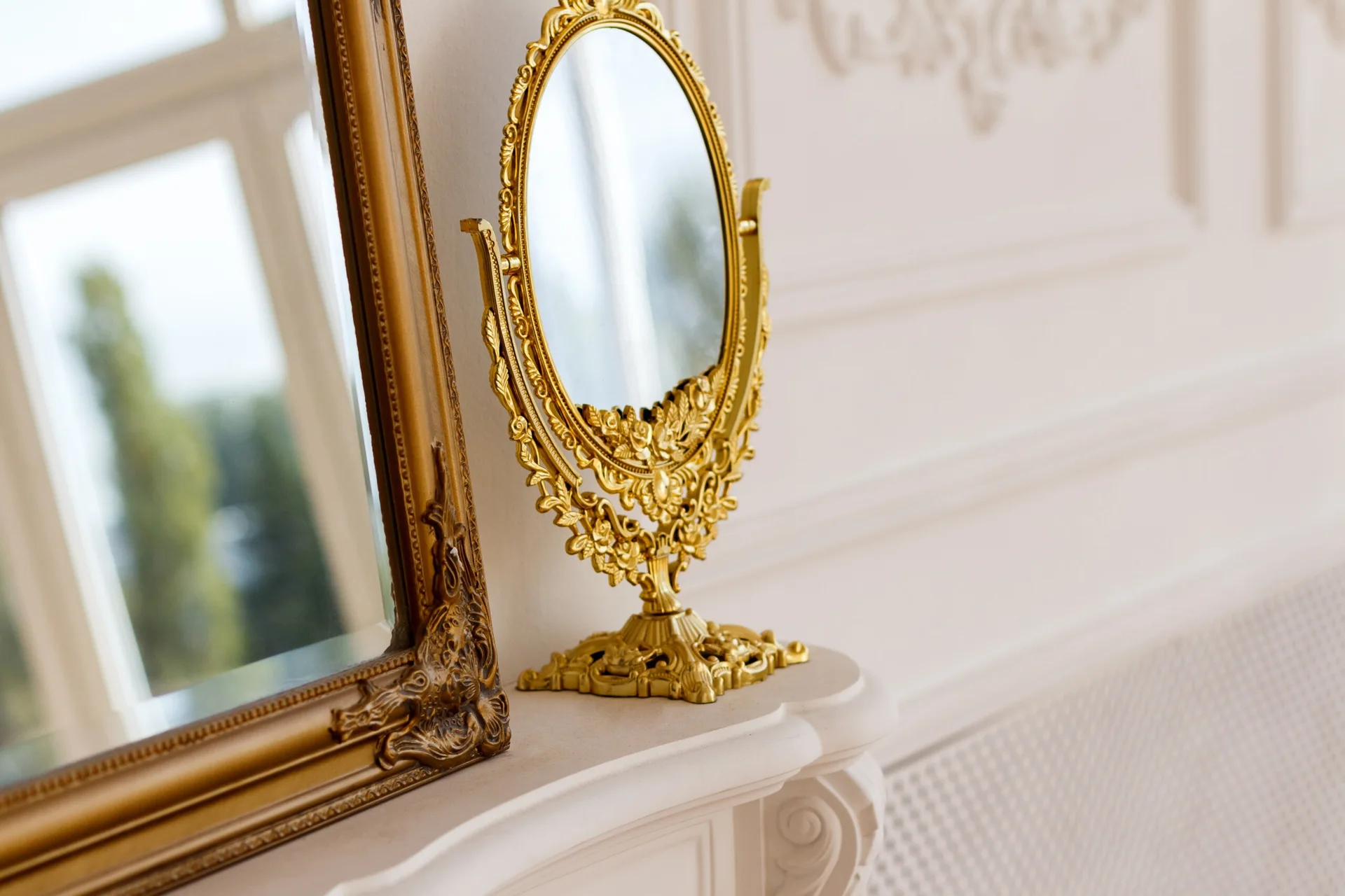 An ornate oval gold tabletop mirror stands on a white mantelpiece next to a larger gold-framed mirror against a wall with decorative molding.