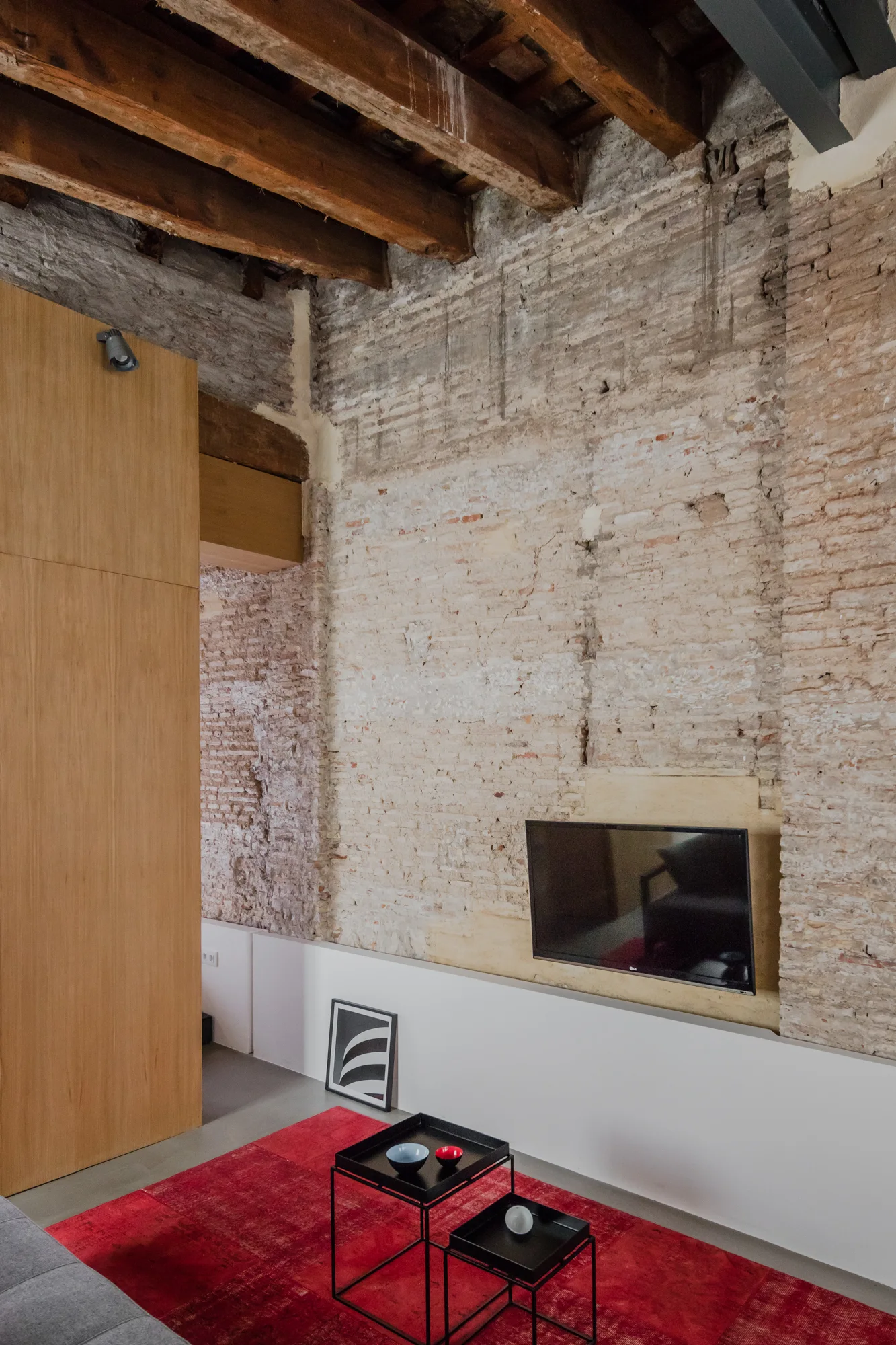 A living room features an exposed brick wall, dark wood ceiling beams, a wall-mounted TV, a red area rug, and black metal coffee tables.