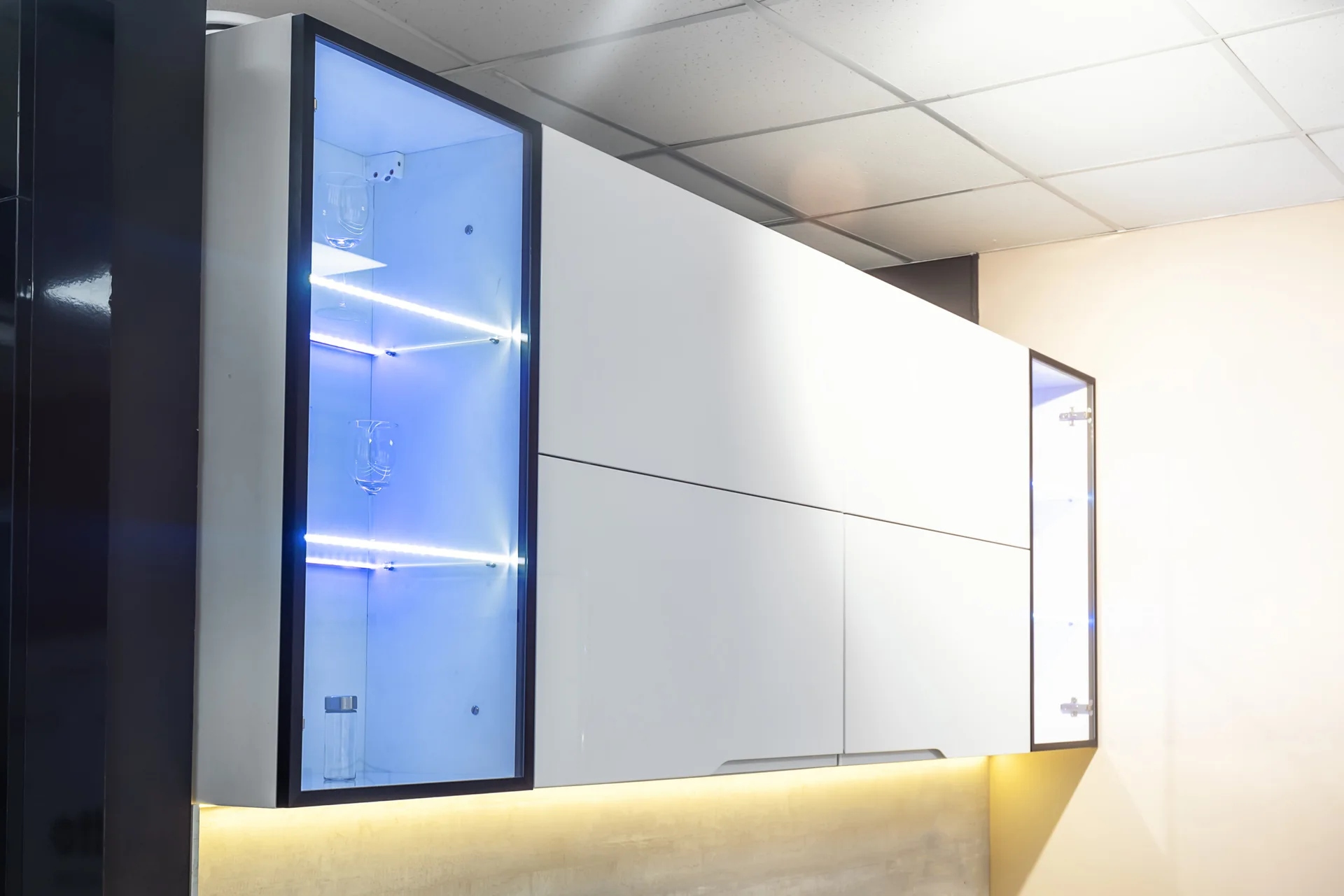 Modern high-gloss white kitchen wall cabinets feature glass-front display sections illuminated with blue LED strip lighting and a warm under-cabinet glow.