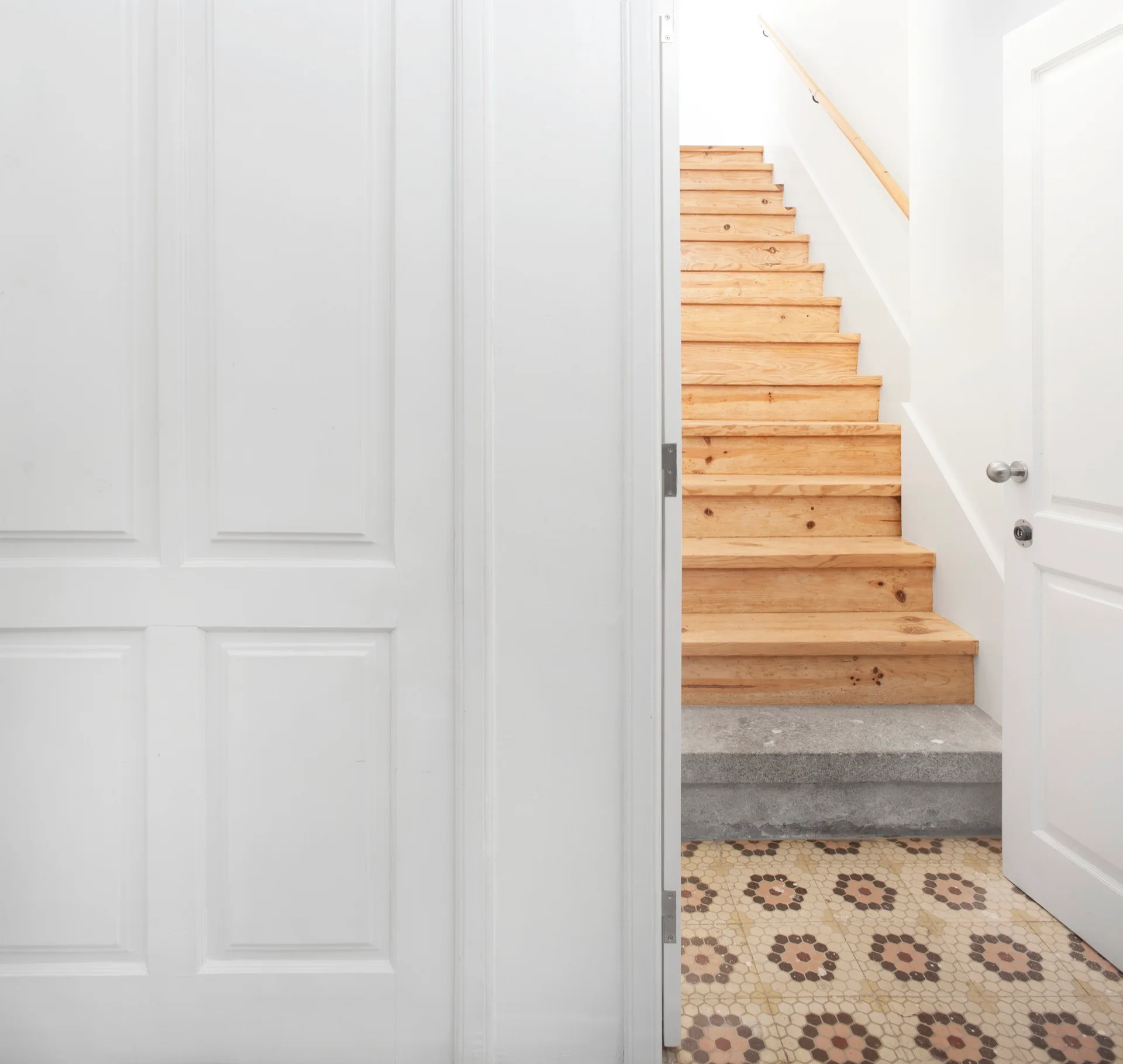 A view through open white doors shows a light wood staircase rising above a patterned hexagonal tile floor and a grey concrete base step.