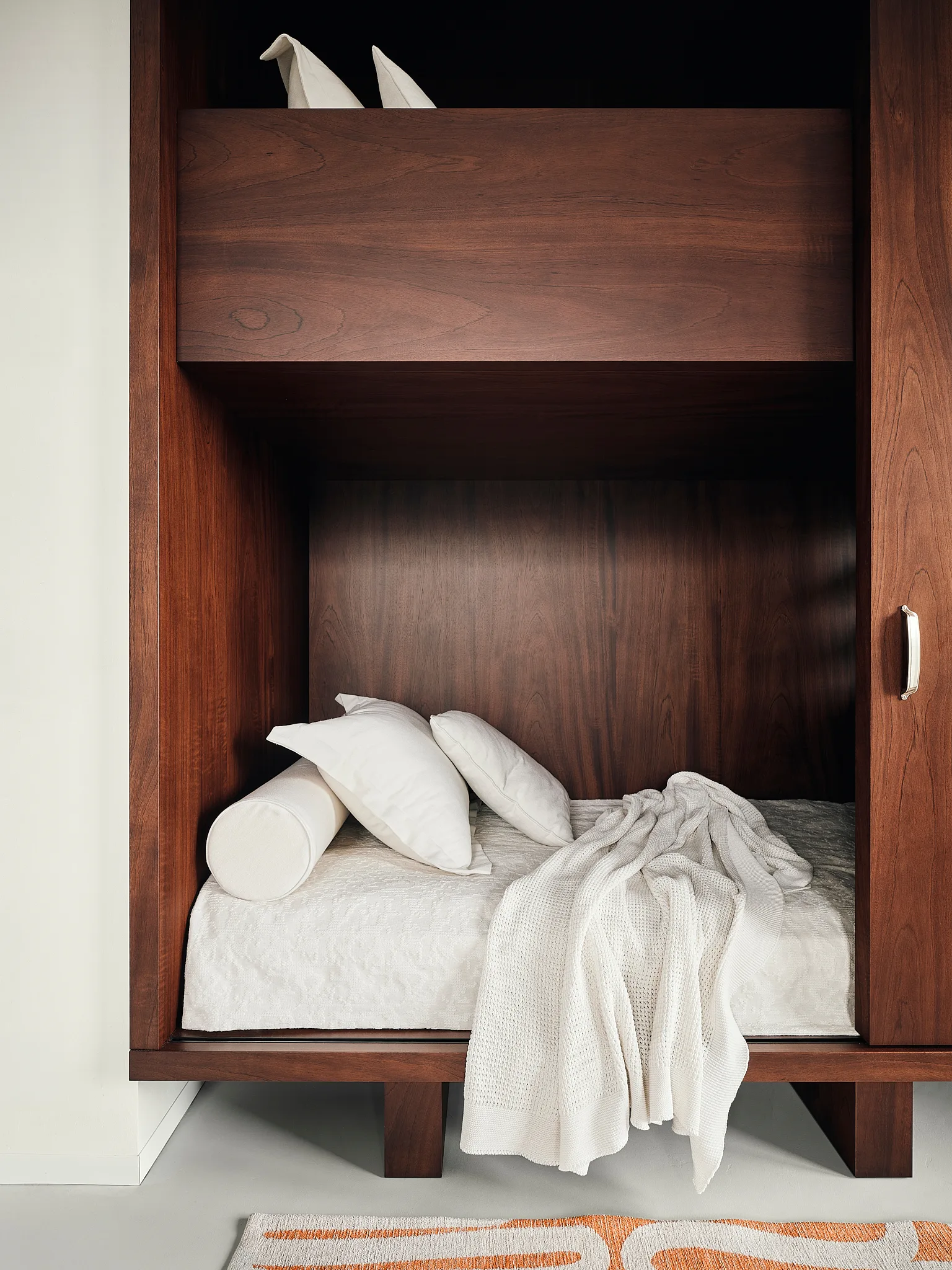 A built-in dark wood bed alcove is tucked into custom walnut cabinetry and dressed in clean white bedding, pillows, and a textured throw blanket.