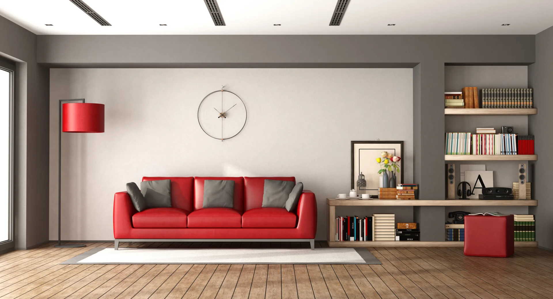 A grey living room with a bright red leather sofa, built-in wooden shelving, a red floor lamp, and a minimalist wall clock.