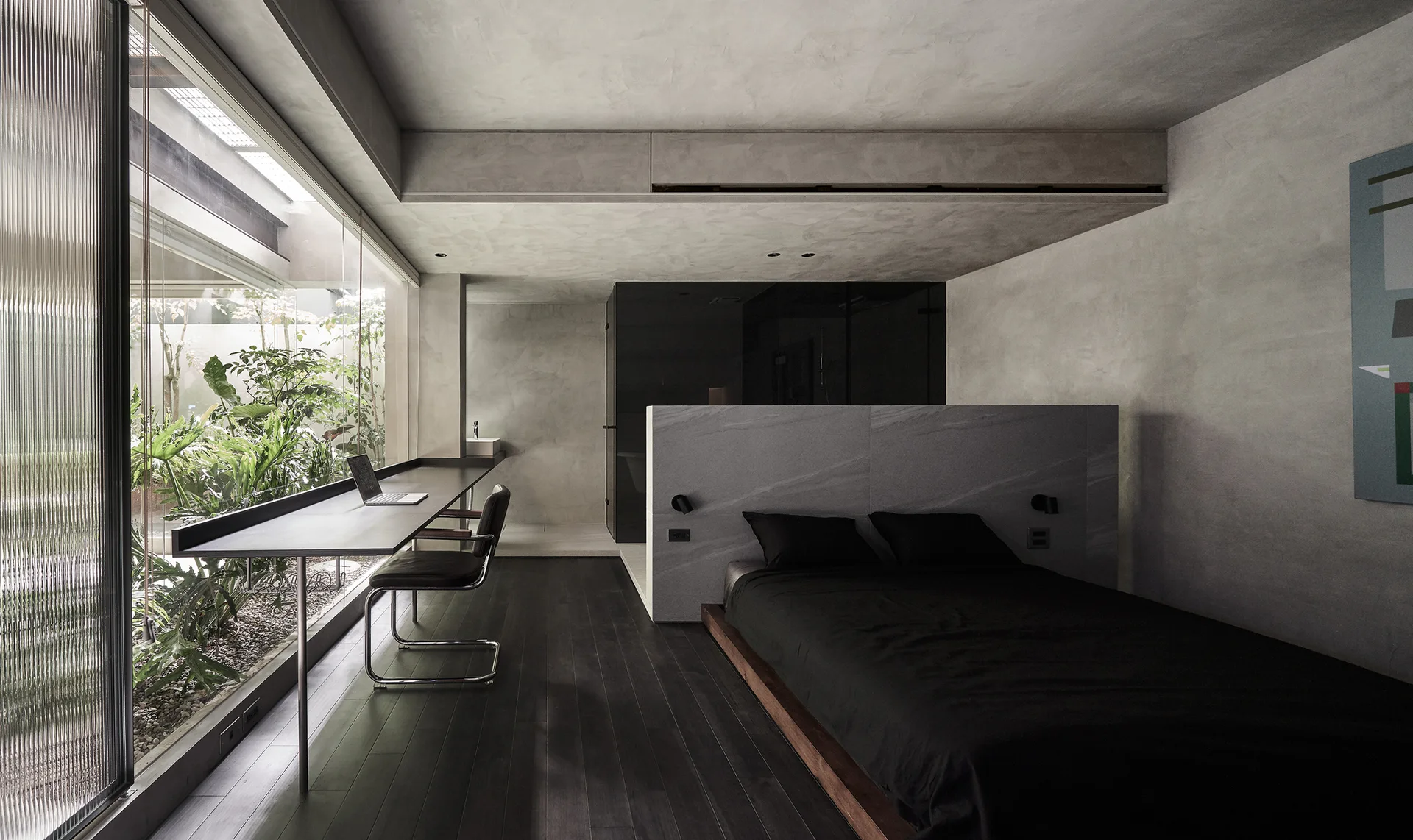 A minimalist concrete bedroom featuring a low platform bed with a stone headboard partition and a long built-in desk facing a garden window.