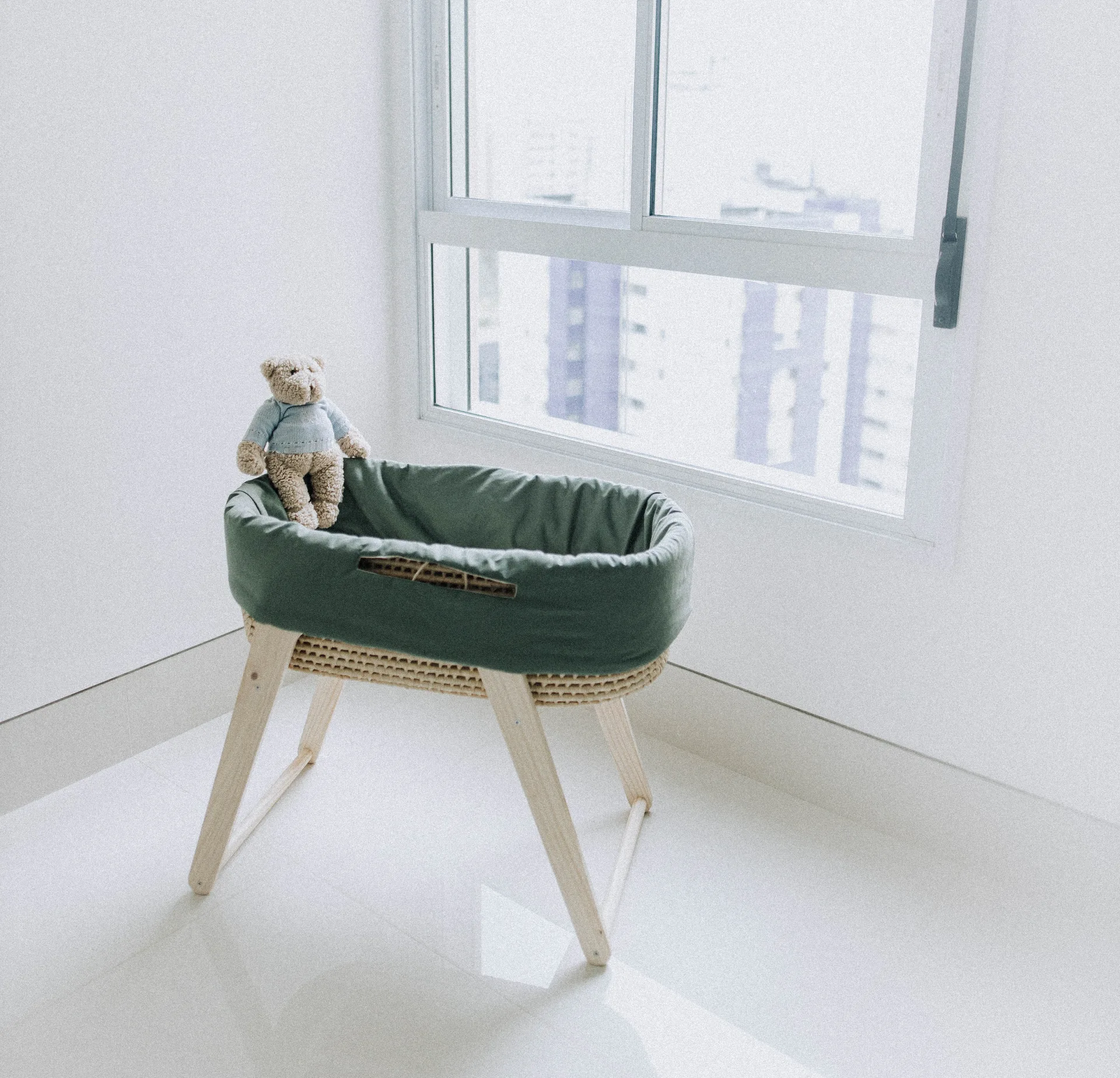 A sage green fabric bassinet on a light wood stand sits next to a large window in a bright minimalist nursery.