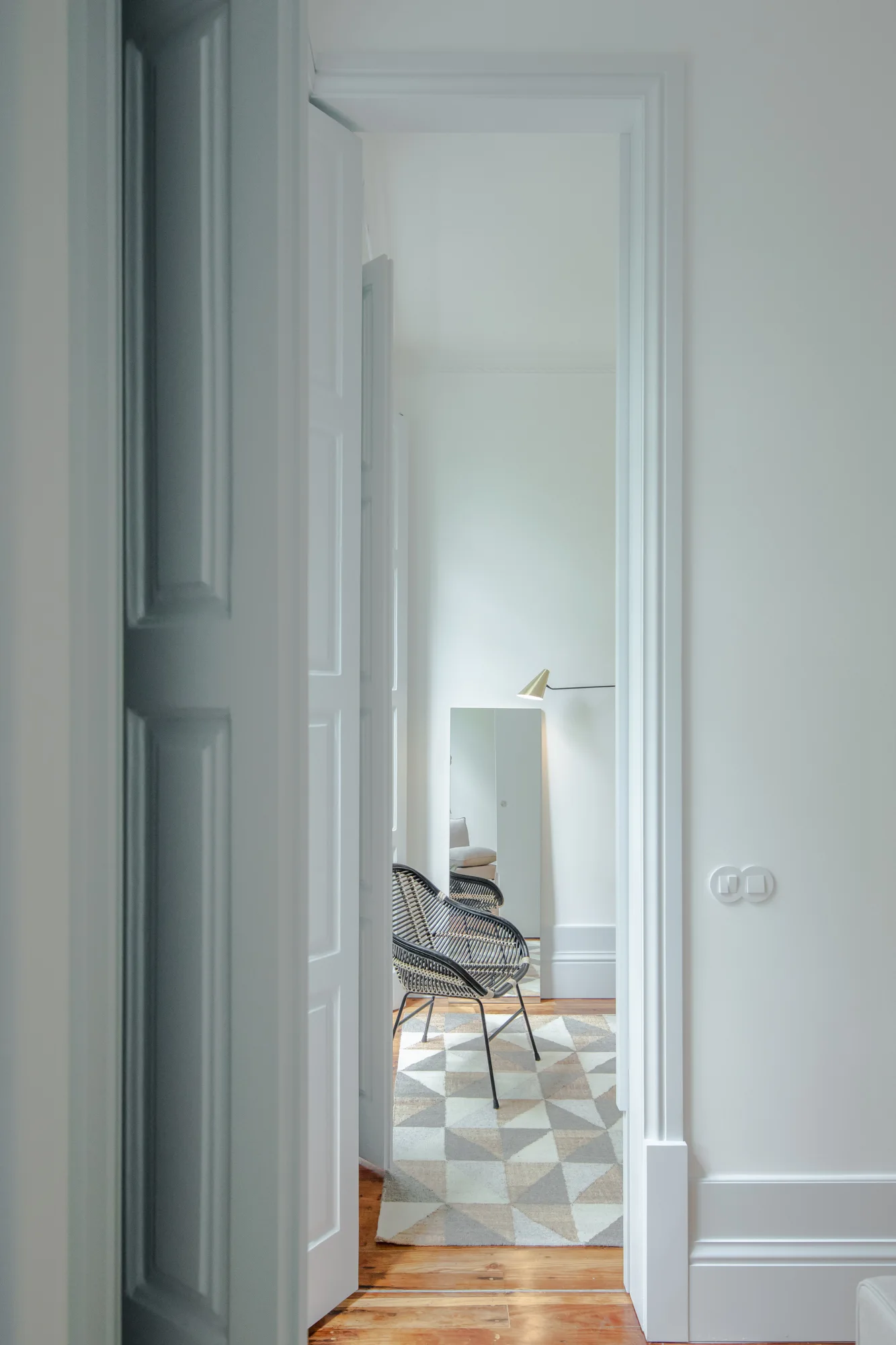 A view through a white doorway into a minimalist room featuring a black woven accent chair, a geometric patterned rug, and a tall floor mirror.