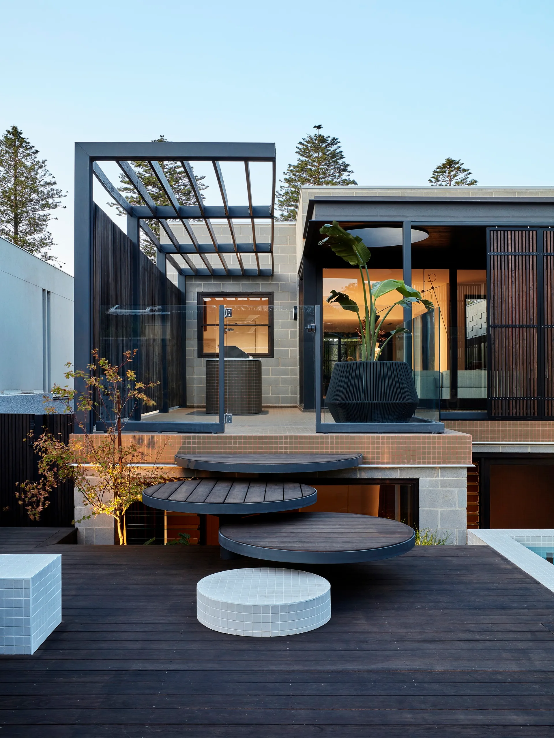 A modern multi-level outdoor deck features circular floating wooden steps connecting to a raised tiled patio with a black pergola and glass railing.