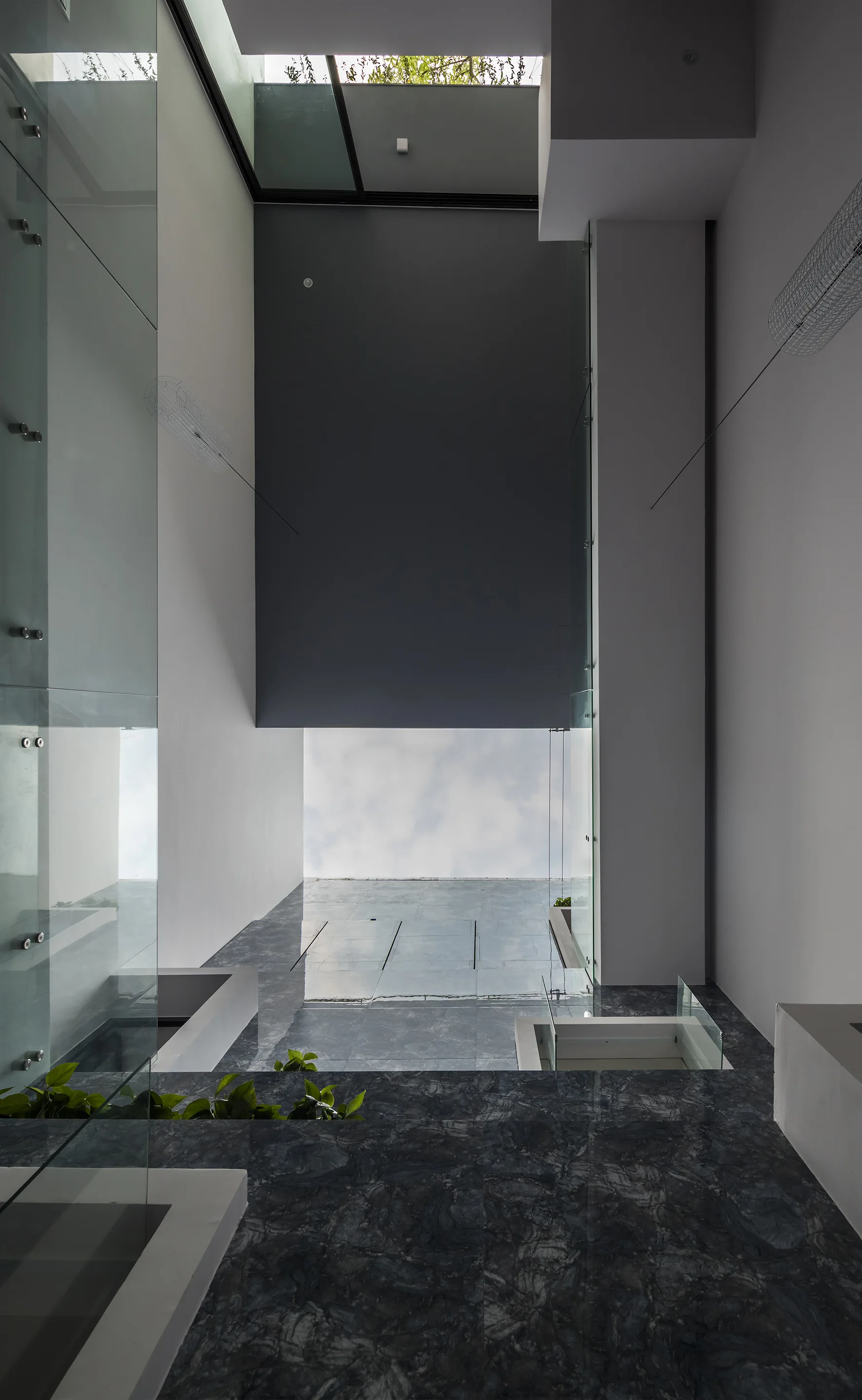 A modern double-height atrium features dark polished marble floors, clean white walls, glass railings, and a large overhead skylight showing a cloudy sky.