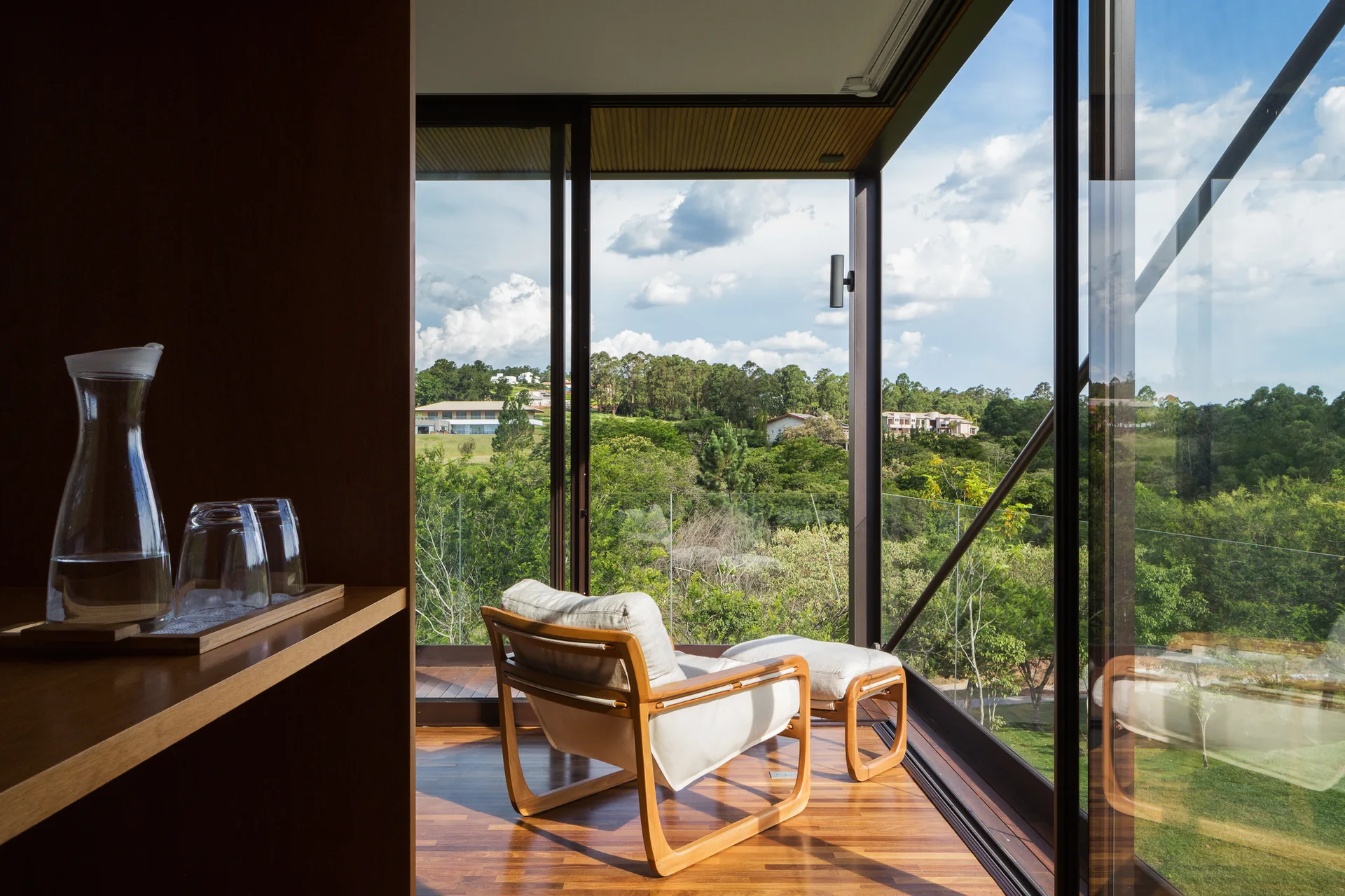 A wooden lounge chair and ottoman face a large panoramic window overlooking a green landscape.